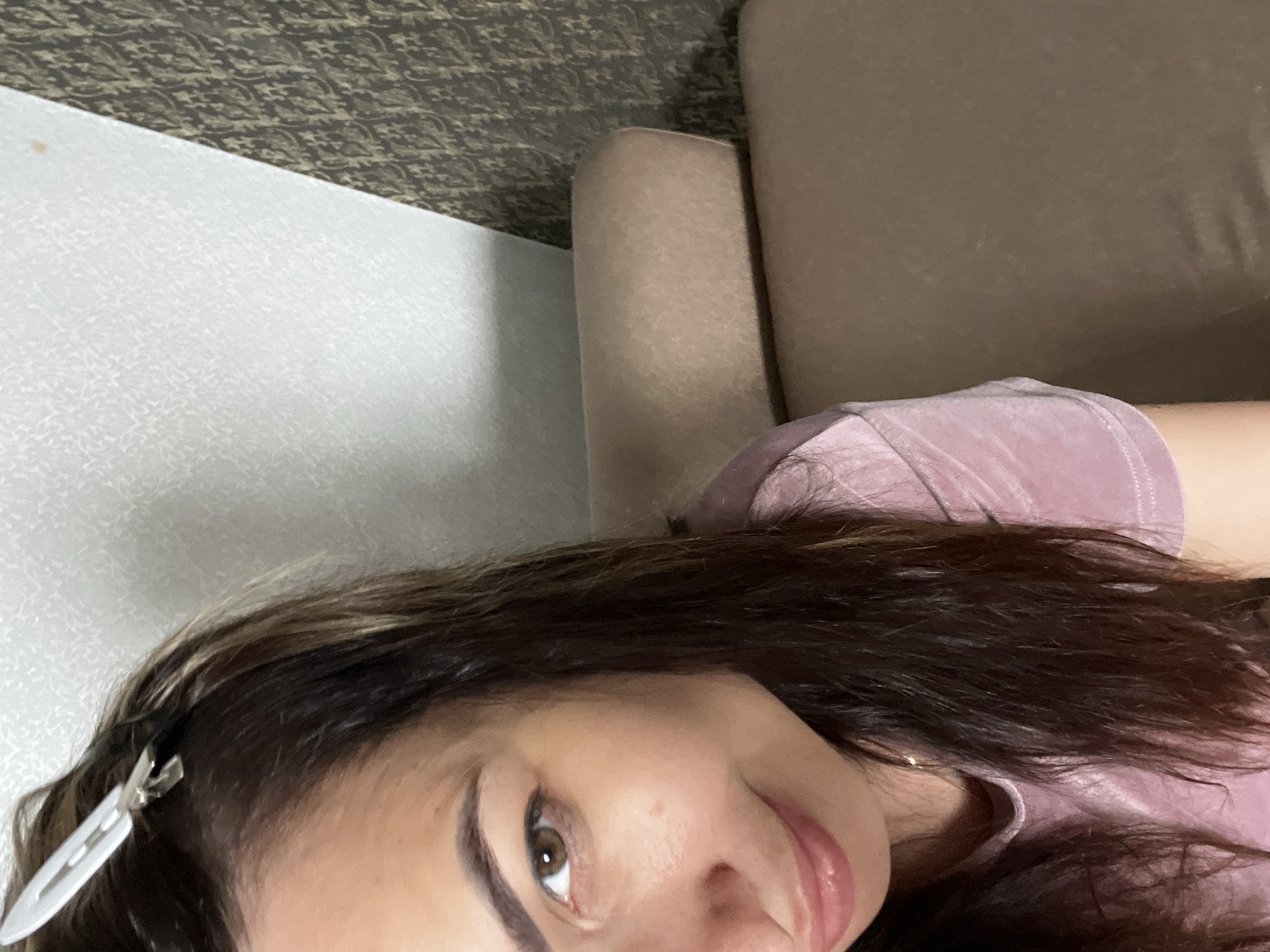 A woman with dark hair and brown eyes is looking at the camera. She is wearing a pink top and has a white hair clip in her hair. She is sitting on a couch in a room. The wall behind her is decorated with wallpaper.