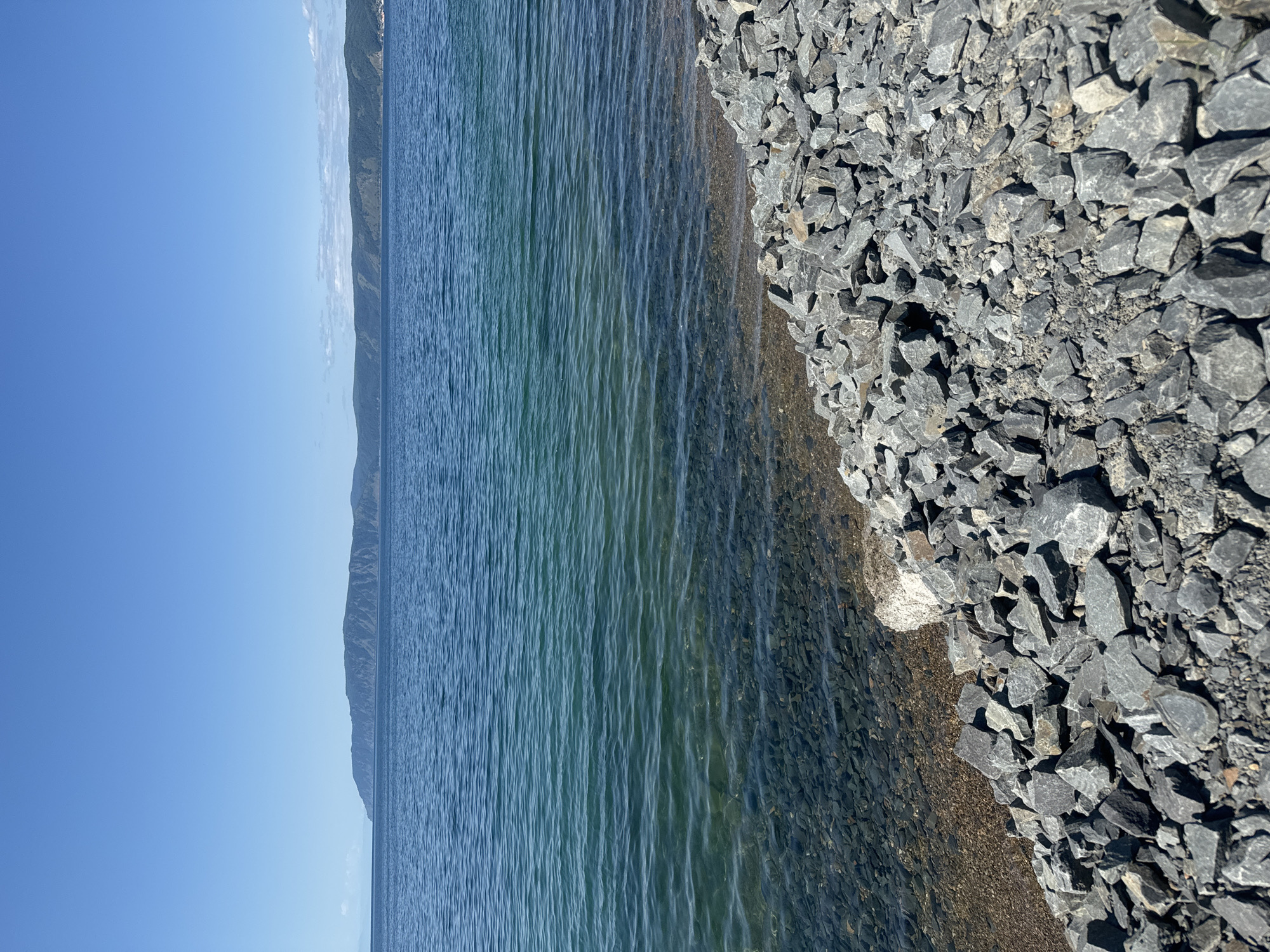 The image depicts a view of the shore of Lake Baikal in Listvyanka, Russia. The foreground is dominated by a rocky embankment composed of numerous gray stones. The water of the lake is clear and exhibits a gradient of blues and greens, with the bottom visible in the shallow areas near the shore, revealing more stones and pebbles. In the middle ground, the expanse of the lake stretches out, with gentle ripples on its surface. The background features a distant shoreline with rolling hills covered in vegetation. The sky above is a clear, bright blue with no clouds visible. The scene is tranquil and natural, with no people or man-made structures other than the implied embankment.