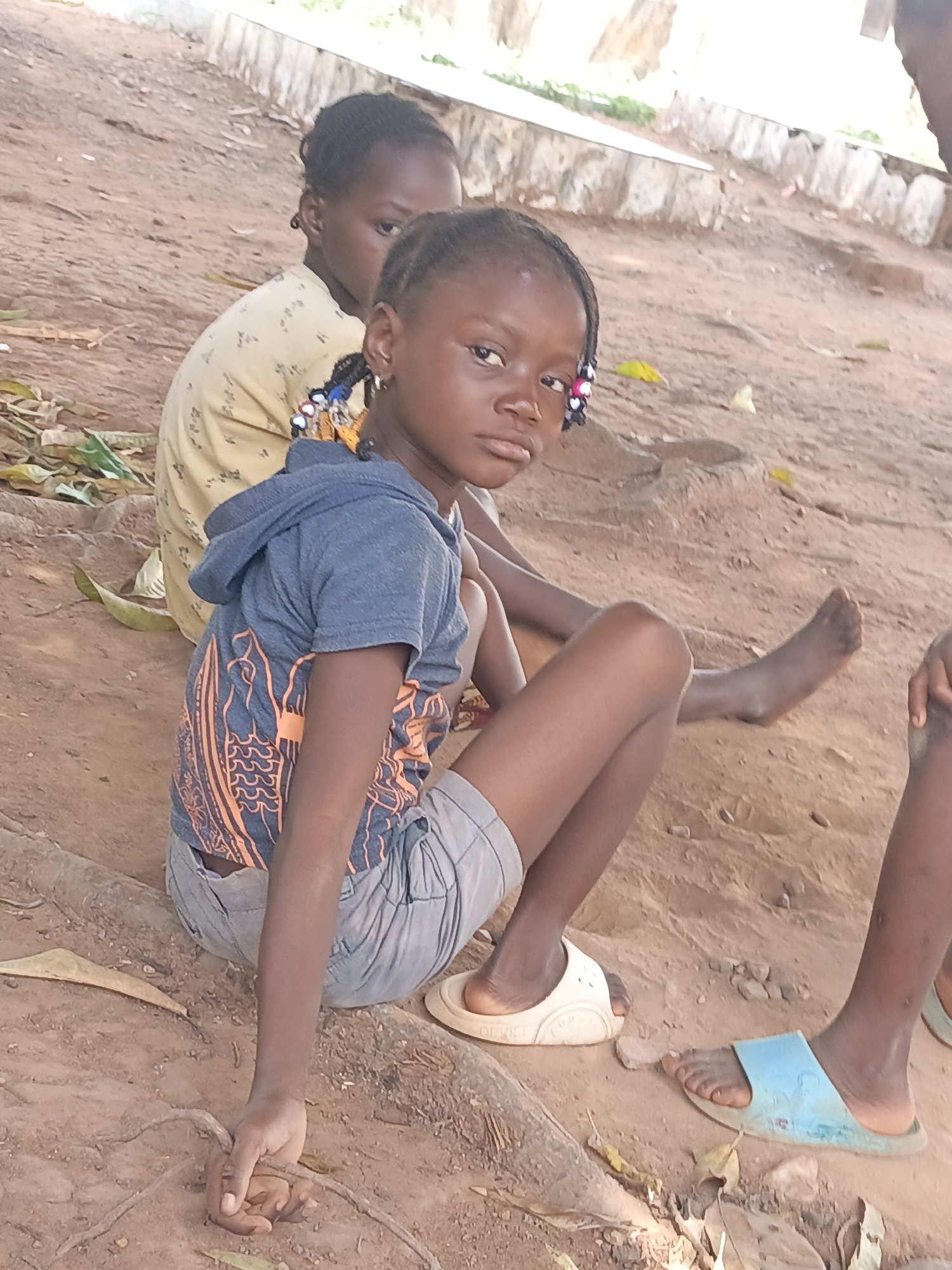 Two young girls sit on the ground in Akwanga, Nigeria. The girl in the foreground wears a blue shirt with an orange design, grey shorts, and a white sandal. She is holding a piece of rope in her hand. The other girl wears a yellow shirt with a small design. Another person's foot, wearing a blue sandal, is visible in the lower right corner of the image. The ground is dirt and leaves. A low stone wall is visible in the background.