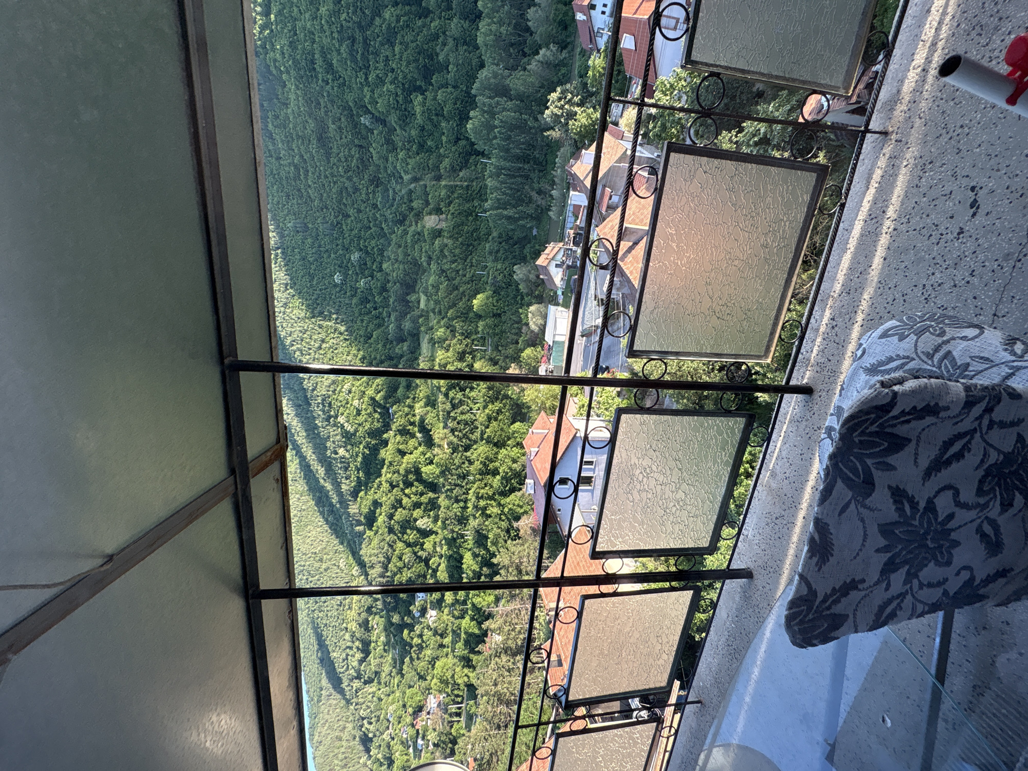 A high-angle exterior shot captures a view from a covered balcony overlooking a village nestled in a lush green valley. The balcony features a metal railing with decorative circular accents and frosted glass panels. A patterned armchair is visible in the foreground on the textured concrete floor of the balcony. Below, a cluster of houses with red-tiled roofs are scattered amongst the trees. The background is dominated by a densely forested mountainside, bathed in the dappled light of what appears to be late afternoon. The scene suggests a peaceful, rural setting in Redina, Bulgaria.