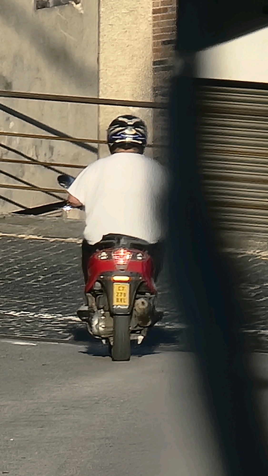 A person wearing a white t-shirt and a helmet is riding a red scooter down a paved street in Sant Joan Les Fonts, Spain. The scooter has a license plate that reads "C7 273 BAL". The person is viewed from behind. The background shows a building with a brick wall and a large metal garage door. There is also a metal railing in the foreground.