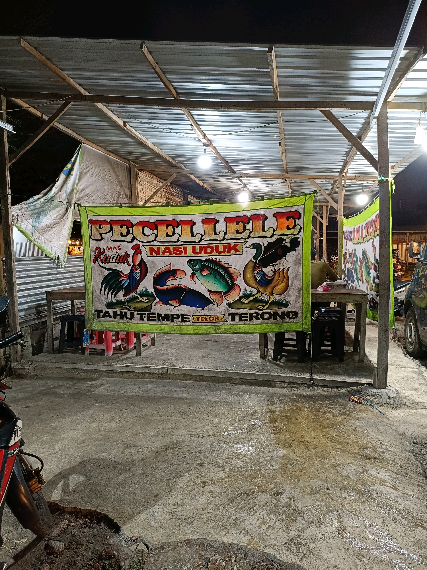 A wide banner displays "PECEL LELE NASI UDUK MAS KEMEK" with colorful illustrations of animals and food items. The banner is hung under a makeshift roof structure supported by wooden beams and corrugated metal. In front of the banner, a wooden table with plastic chairs is set up on a concrete floor. A person is seated at a table behind the banner. To the left, a motorcycle is partially visible. The scene appears to be an outdoor food stall in Maguwoharjo, Indonesia, possibly at night, given the artificial lighting.