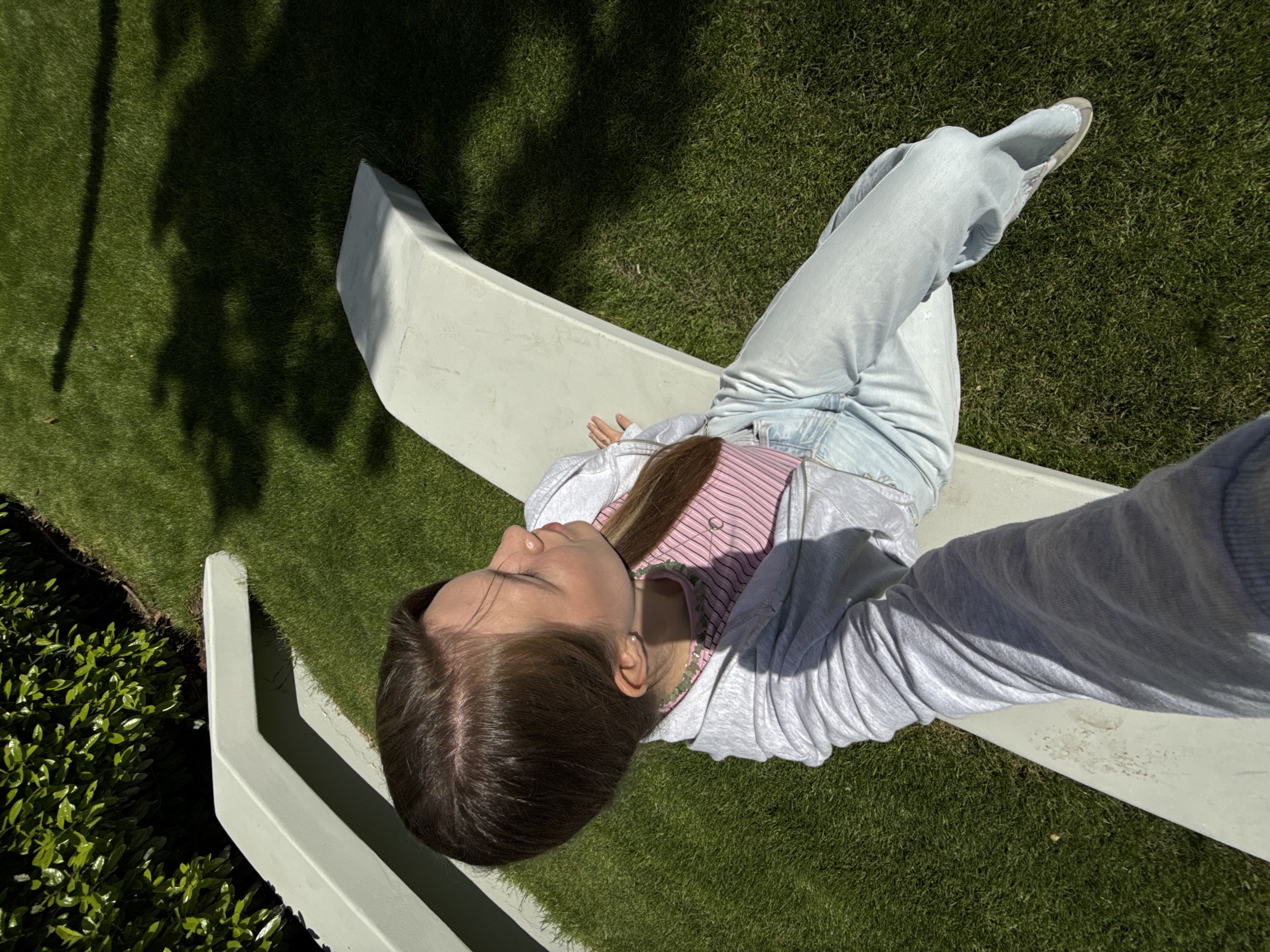 A young woman lies on a concrete bench in what appears to be an outdoor setting. She is wearing light blue jeans, a pink striped shirt, and a light gray zip-up hoodie. She is lying on her back with her head near the edge of the bench. The environment includes green grass, both in direct sunlight and shadow, and some greenery near the edge of the frame on the left side. The bench is a light gray, possibly concrete or painted material. The scene appears to be outside, and the high angle suggests the photo was taken from above, possibly a selfie or from an elevated position. The location is unknown. The setting is outdoor, and the high angle suggests the photo was taken from above. The location of the image is not available.