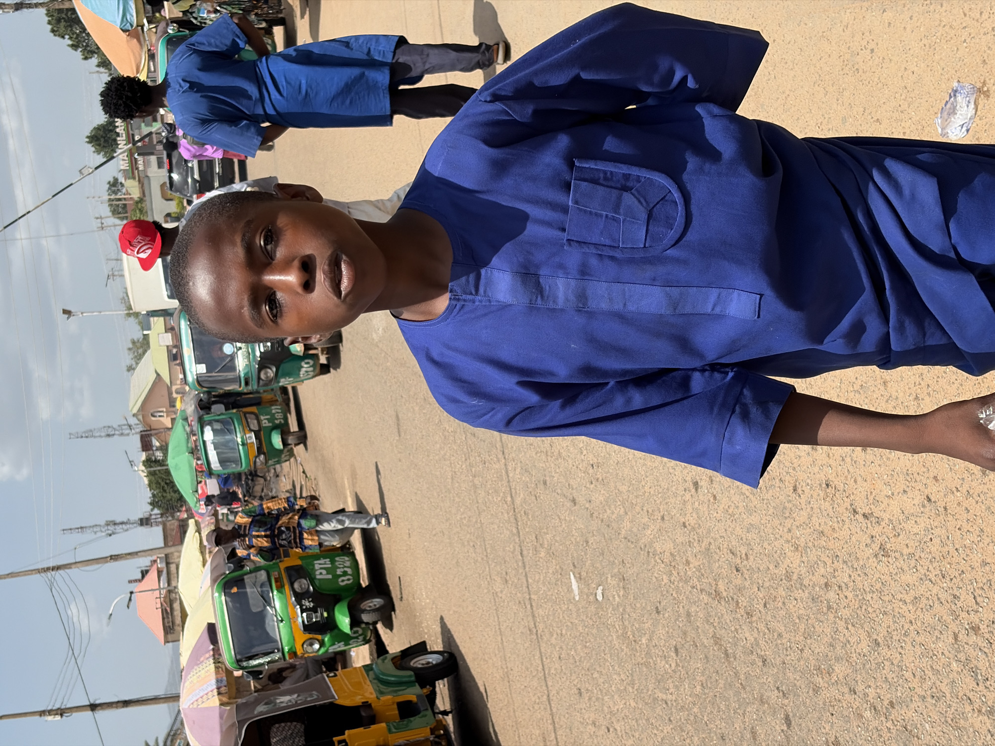 This image captures a vibrant street scene in Jos, Nigeria, under bright daylight.
In the foreground, a young person with dark skin, likely male, looks directly at the camera with a neutral expression. He is dressed in a royal blue, long-sleeved kaftan-style tunic, featuring a distinctive, stylized pocket design on the left chest.
The background reveals a bustling urban environment. Several colorful tricycles, commonly known as Keke Napep, are visible along the paved road. These vehicles are primarily green and yellow, and text on their surfaces includes "PTA 8320" and a partially visible "5871." Further back, another individual wearing a similar blue kaftan is seen from behind, walking, and a person in a red cap is partially visible behind the main subject.
The setting includes various buildings with light-colored roofs in the distance, and utility poles with wires crisscross against a clear, bright sky. Strong shadows cast by the poles and people indicate a sunny day, likely mid-morning to afternoon. A white sign with the word "MA'ANLAN" is noticeable in the mid-ground, suggesting a local business. The ground is a light-colored paved surface, with scattered small pieces of white litter. The overall scene depicts everyday life and transportation in a Nigerian city street.