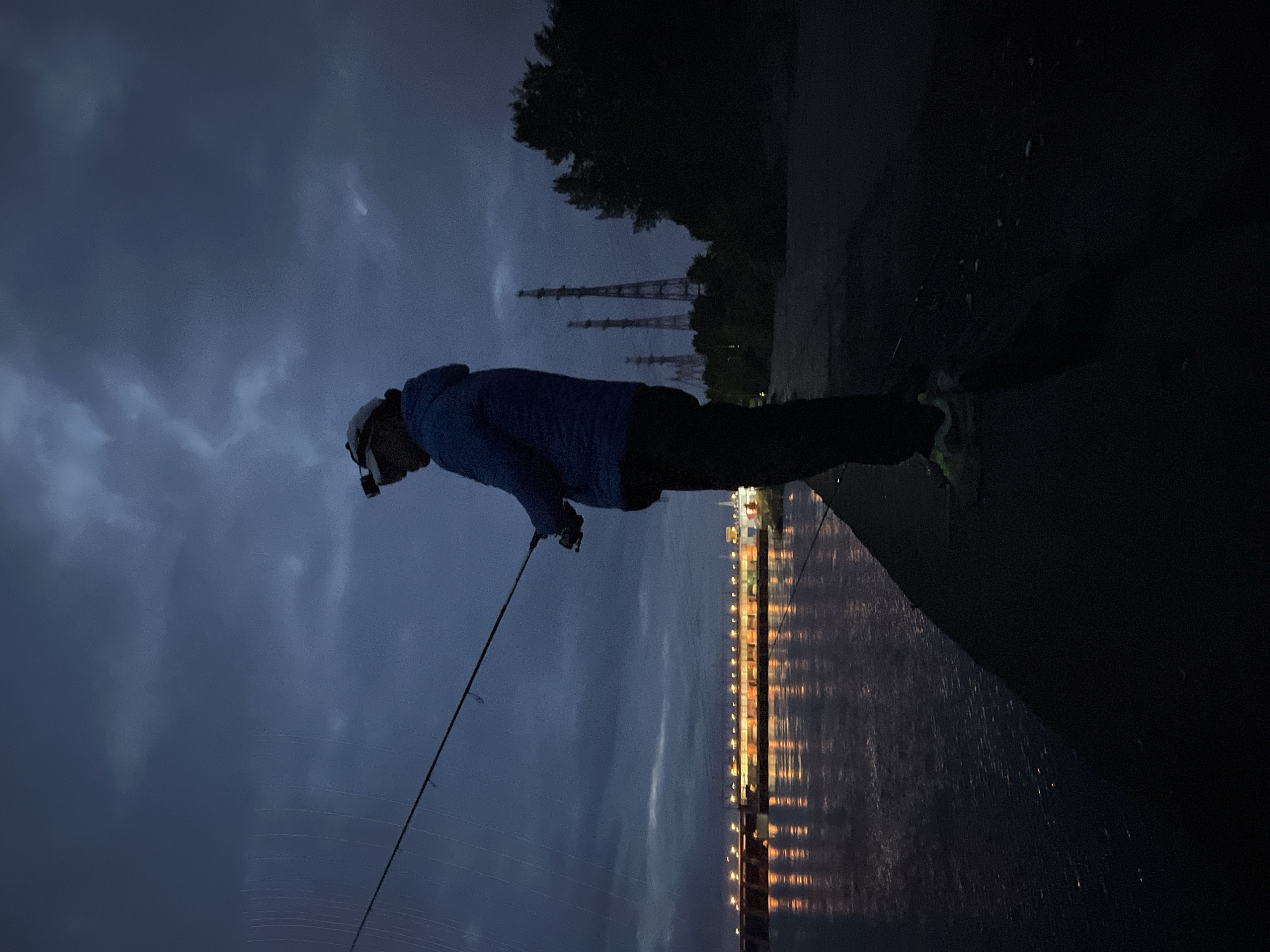 The image depicts a pre-dawn or dusk scene in Chaikovsky, Russia, specifically along the Kama River near the Votkinsk Hydroelectric Power Plant. A lone individual is positioned in profile, facing left, standing on a dark, angled concrete or asphalt embankment. The person is an adult, wearing a light-colored baseball cap with a dark headlamp affixed to the front, a blue hooded jacket, dark trousers, and light-colored athletic shoes. They are holding a fishing rod with a spinning reel, angling into the body of water.
To the left of the individual is the dark, rippled surface of the water, reflecting numerous elongated, bright orange and white lights from a large structure on the far bank. This structure is the Votkinsk Hydroelectric Power Plant dam, characterized by its long expanse of illuminated buildings and operational sections.
To the right of the individual, extending into the midground, are several tall, dark, lattice-like structures, indicative of power transmission towers or industrial masts. These are silhouetted against the dark sky. Behind these towers and further to the right, a dense mass of dark trees forms a silhouette. The sky above is overcast with dark blue-grey clouds, suggesting heavy cloud cover or low light conditions.