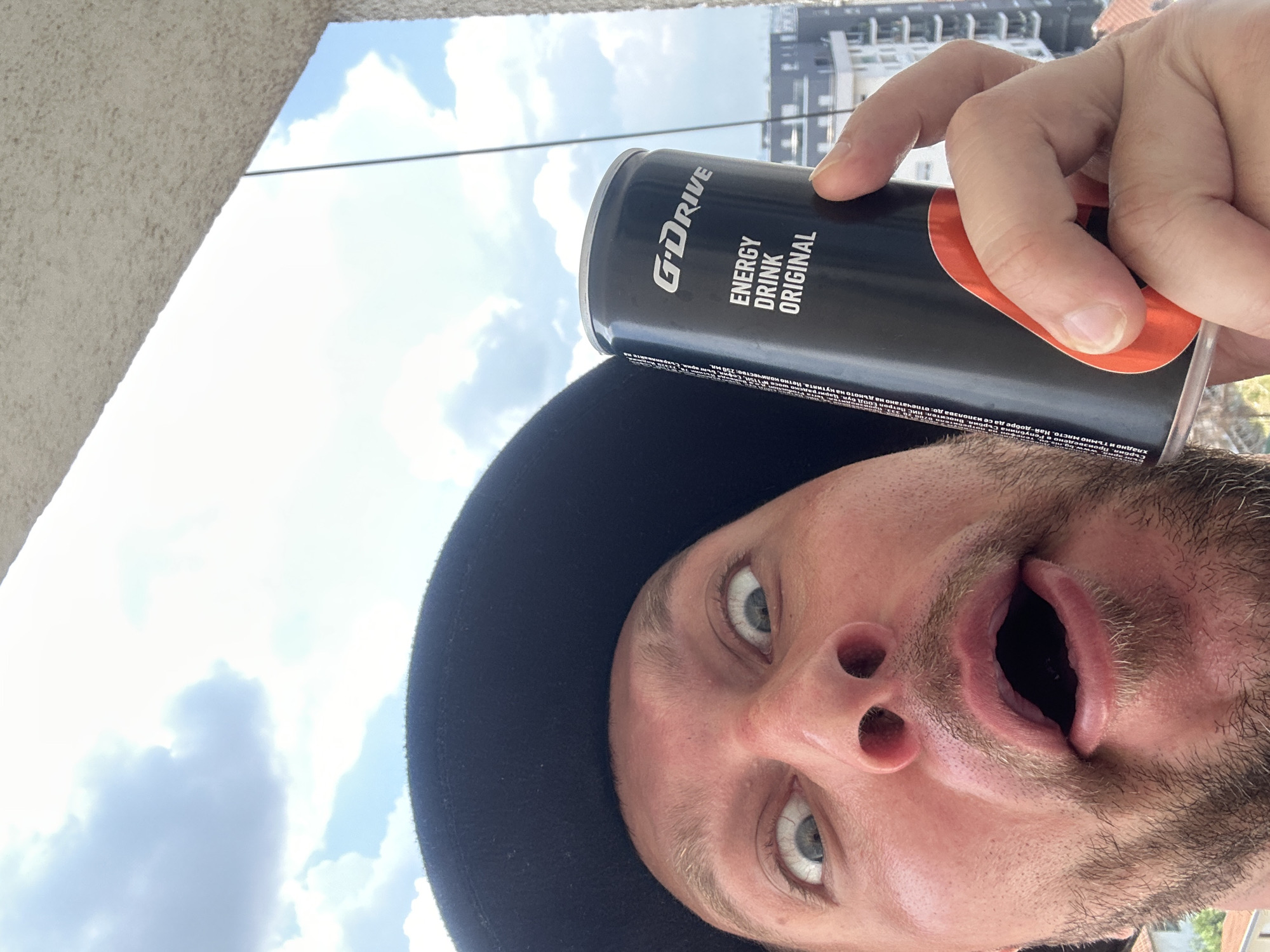 A man wearing a black hat is holding up a black can of "G'DRIVE ENERGY DRINK ORIGINAL" to the camera, making a surprised face with wide eyes and an open mouth. The background shows a cloudy sky and buildings. This appears to be a selfie taken outdoors, possibly on a balcony in Zvezdara Urban Municipality, Serbia.