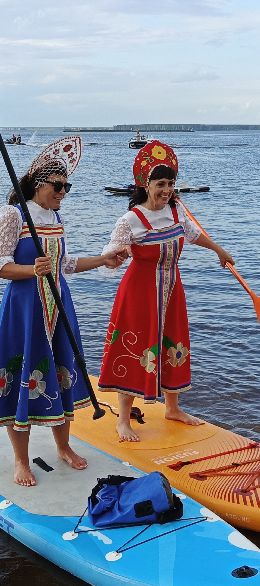 Two women, dressed in traditional Russian attire and headdresses, are standing on two paddleboards on a body of water. The woman on the left wears a blue dress and a white headpiece adorned with red and white embroidery. She holds a paddle and wears sunglasses. The woman on the right wears a red dress and a red headdress with yellow flowers. She holds an orange paddle and is smiling. Both women are holding hands and appear to be interacting. The paddleboards are orange and blue and are next to each other. A blue waterproof bag is visible on the blue paddleboard. In the background, the sky is partly cloudy and the water is calm. Several boats and jet skis are visible in the distance, suggesting a recreational area. The location is the Kama River in the city of Chaikovsky, Russia.