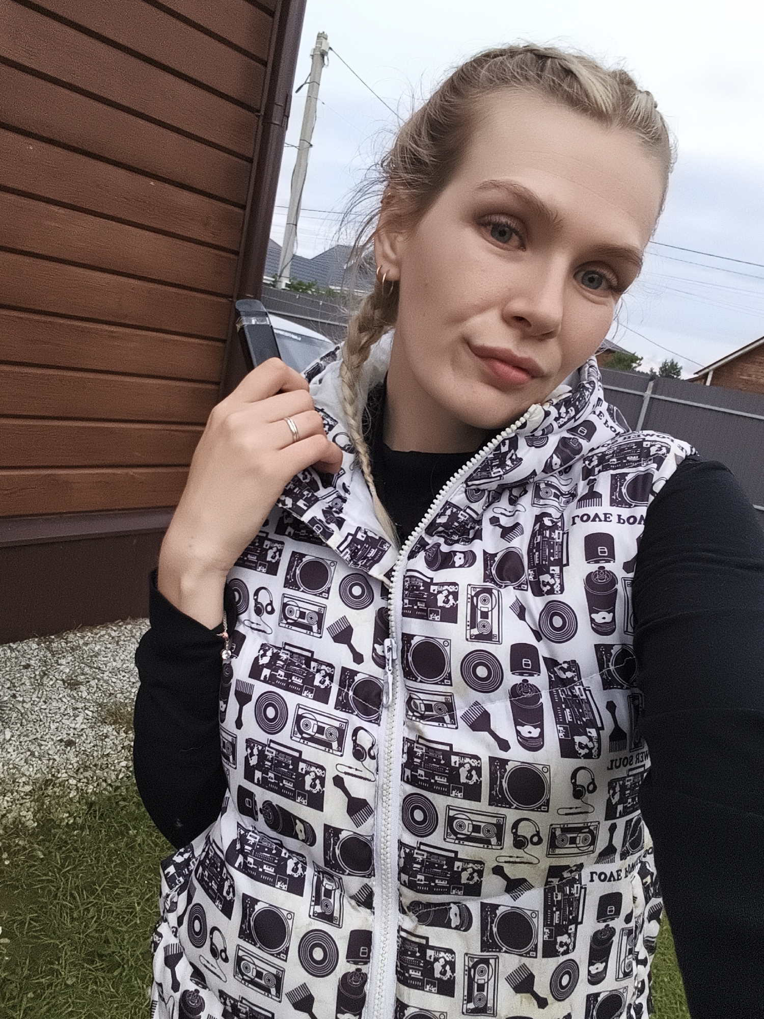 A young woman with blonde hair braided into two pigtails is standing outdoors, taking a selfie. She is wearing a black long-sleeved shirt and a white vest with a black pattern of music-related imagery, including boomboxes, headphones, vinyl records, and spray paint cans. A silver ring is visible on her left hand, which is resting on her collarbone. She is wearing small hoop earrings. Her makeup includes winged eyeliner and lip gloss.
The background shows a brown wooden building, a grey fence, and some greenery, including grass and what appears to be gravel. A utility pole with wires is visible in the distance. The sky is overcast, suggesting a cloudy day. The image appears to be taken during daylight hours. The text on the vest, when reversed, reads "GOLE BOA" and "JUOS RAW," which are likely design elements rather than descriptive text. The overall impression is casual and relaxed.
