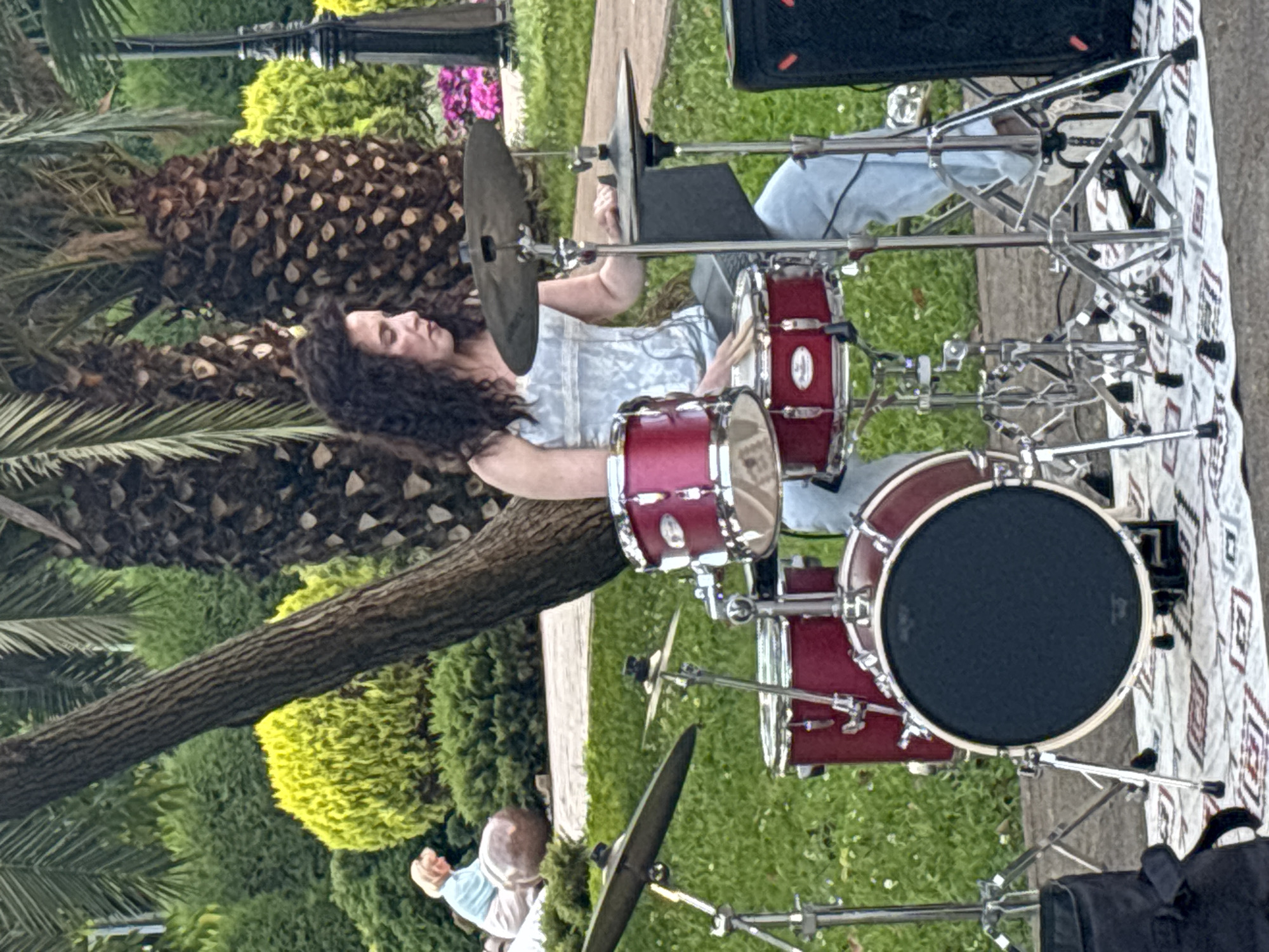 An outdoor shot captures a woman playing a red drum set. She has dark, curly hair and is wearing a light-colored, sleeveless top and light blue pants. She is seated behind the drum set, which includes a snare drum, tom-toms, a bass drum, and cymbals. Her hands are on drumsticks, poised to strike the drums. In the background, palm trees, green bushes, and a person sitting on the ground are visible. The setting appears to be a park or garden, possibly in Sochi, Russia, given the presence of palm trees and the general landscape.