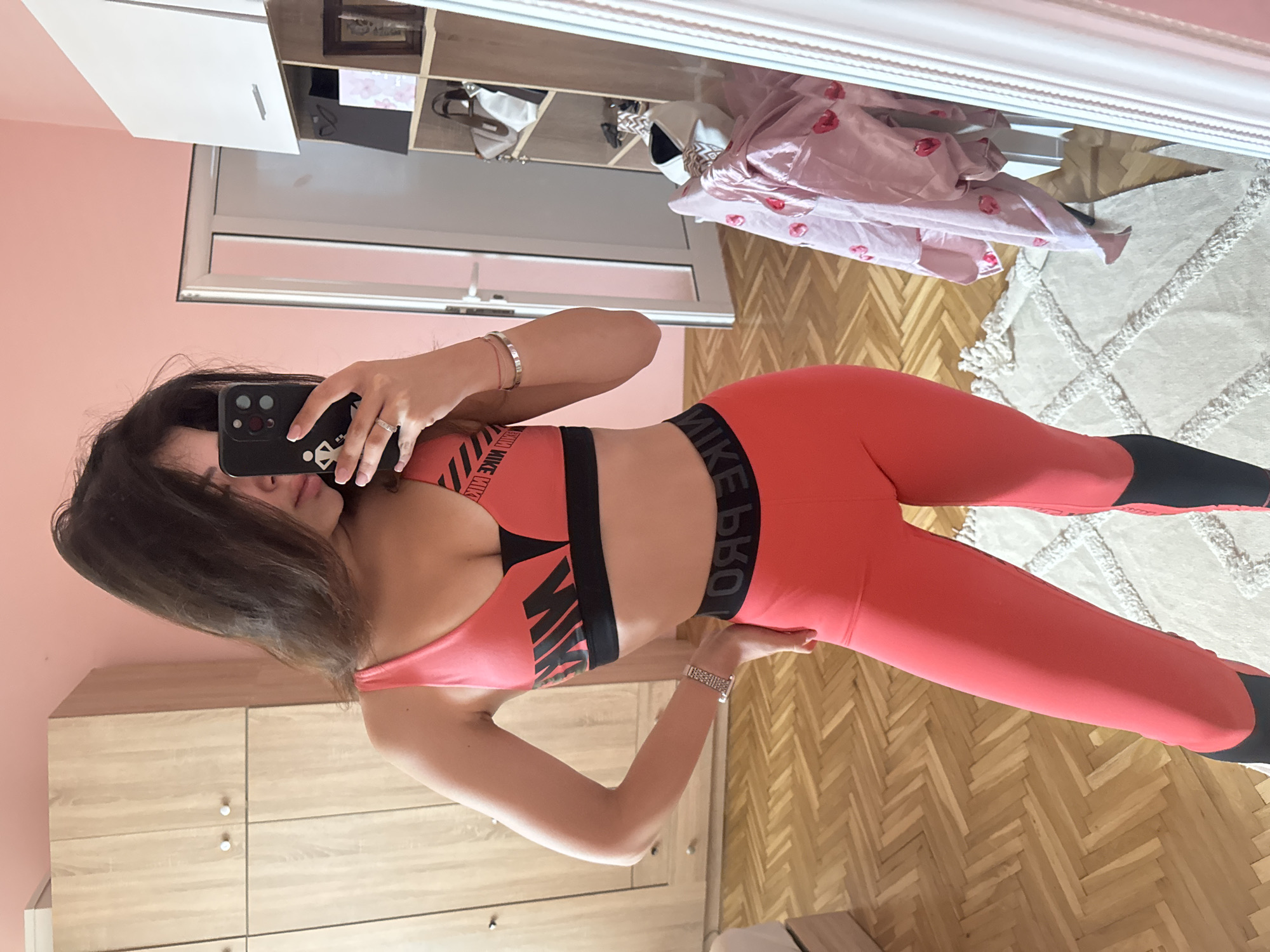 A young woman with dark hair is taking a mirror selfie in a bedroom. She is wearing a coral-colored sports bra and matching leggings with black accents. The room has light pink walls, light wood cabinets, and a parquet floor. In the reflection, a closet, a shelf unit with various items, and a pink satin robe with heart patterns are visible. The woman is standing on a white rug with a textured pattern. The city of Plovdiv, Bulgaria, is not mentioned or indicated in the image.