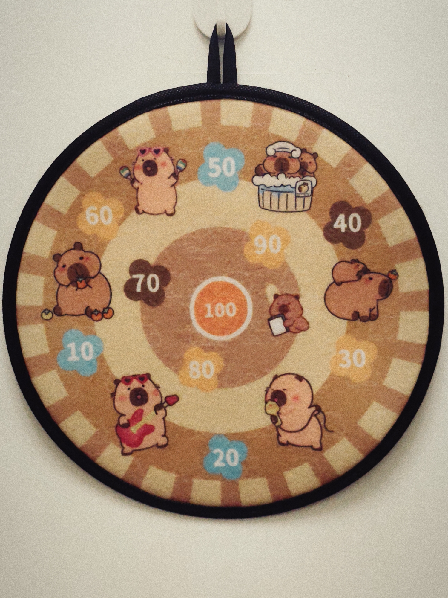 This is a close-up, eye-level shot of a circular dartboard with a cartoonish design. The dartboard is hanging on a plain white wall. The design features a tan background with brown radial stripes and a checkered pattern. Around the outer edge, there are cartoon drawings of capybara-like creatures in various poses, each associated with a number. These numbers are: 50, 40, 30, 20, 10, 60, 70, 80, and 90, arranged in a circular pattern. In the center of the dartboard is a large orange circle with the number 100 inside. The capybara characters are depicted with pinkish-tan skin and dark brown eyes. Some of the characters are wearing accessories like sunglasses or holding musical instruments. The dartboard has a black border and a black loop at the top for hanging. The overall lighting is even, with no harsh shadows.