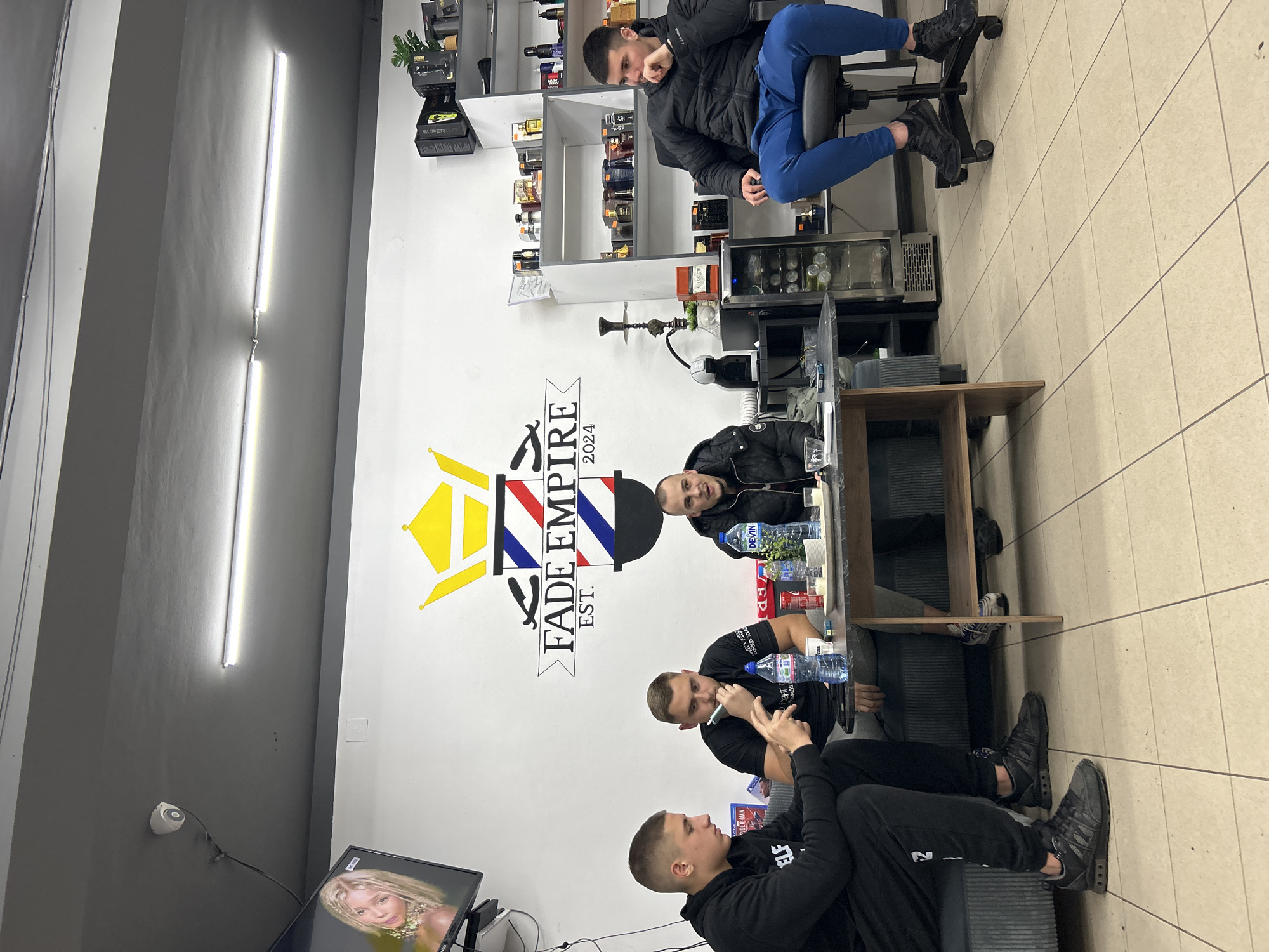 The image shows an interior scene, possibly a barbershop, in Buhovo, Bulgaria. Four men are present: one sits on a swivel chair, wearing a black jacket and blue pants, while three are seated around a table. On the wall, a large logo reads "Fade Empire Est. 2024". A hookah sits on a shelf behind the table. The table has water bottles and other items. A television is visible in the lower left corner, displaying an image of a woman. Fluorescent lights are on the ceiling. The walls are painted white and gray. The floor is covered with square tiles. A security camera is mounted on the wall near the television.