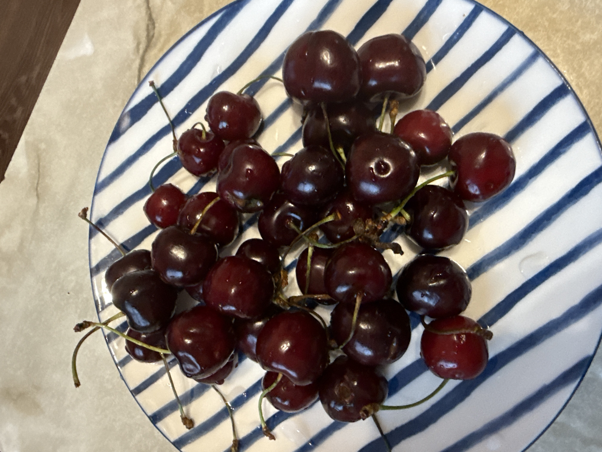 A pile of dark red cherries with stems is arranged on a white plate with blue stripes. The cherries are glossy and some have water droplets on them. The plate is on a light-colored, speckled surface. The scene is lit from above, casting shadows on the cherries and the plate.