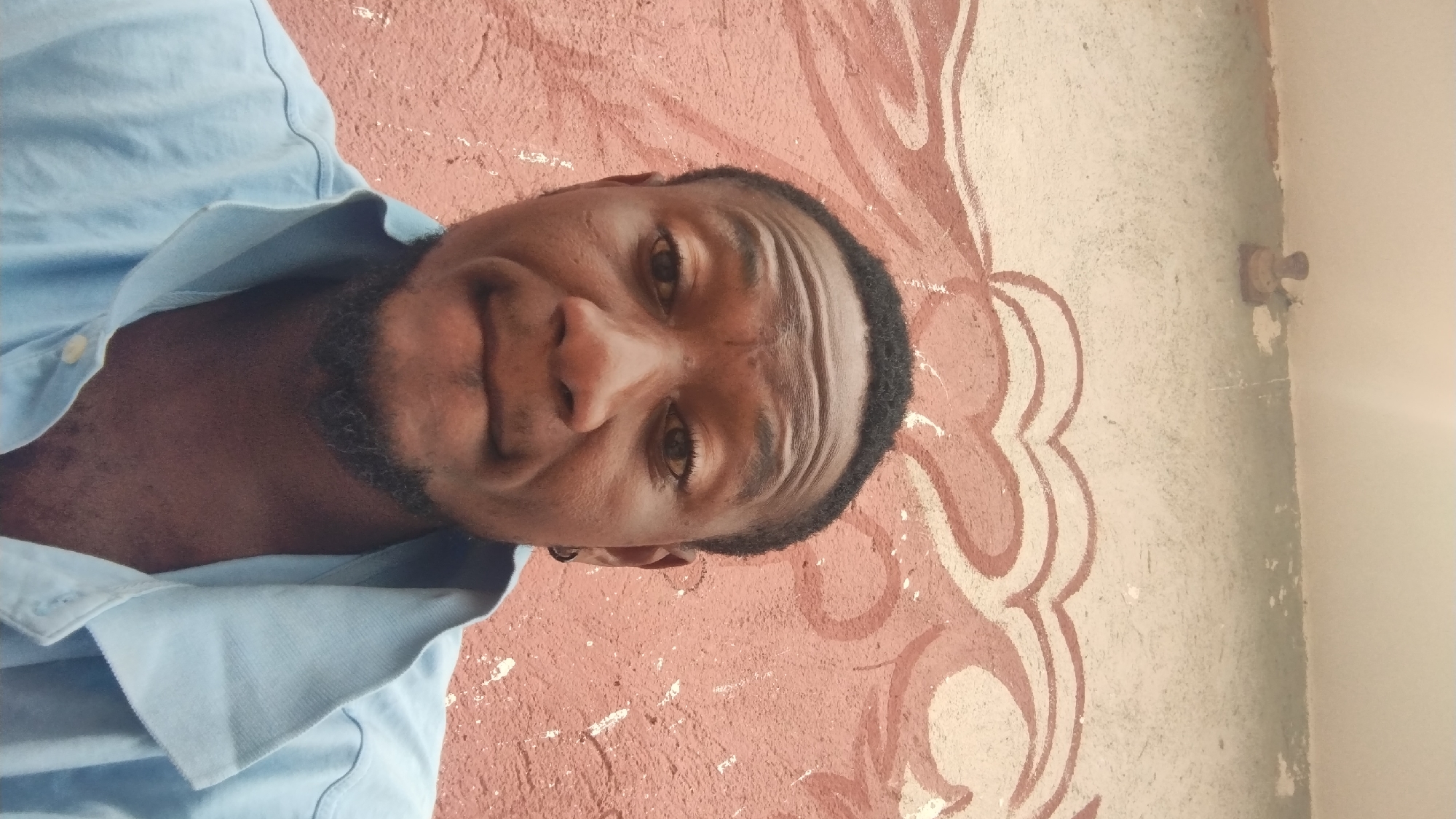 The image displays a close-up portrait of a man, rotated 90 degrees counter-clockwise from its correct orientation. The subject is an adult man with dark skin, short dark hair, and a neatly trimmed dark beard. He is wearing a light blue collared shirt. A small dark earring is visible in his left ear. His facial expression is neutral, possibly hinting at a slight smirk, and his forehead shows several horizontal creases. He is looking directly at the camera.
The background consists of a textured wall, painted with a decorative pattern. The dominant colors of the wall are a rustic reddish-brown and an off-white or light grey. The reddish-brown section features a repeating, lighter-colored design, possibly abstract or stylized floral motifs. To the right, a plainer off-white wall section is visible, and in the upper right corner, a small, simple light fixture or bulb holder is mounted. The overall texture of the wall suggests it is an older or weathered surface. The lighting appears to be natural, indicating daytime. The scene is set against a wall with local artistry, providing a contextual clue for the location, Jos, Nigeria. No visible text is present in the image.