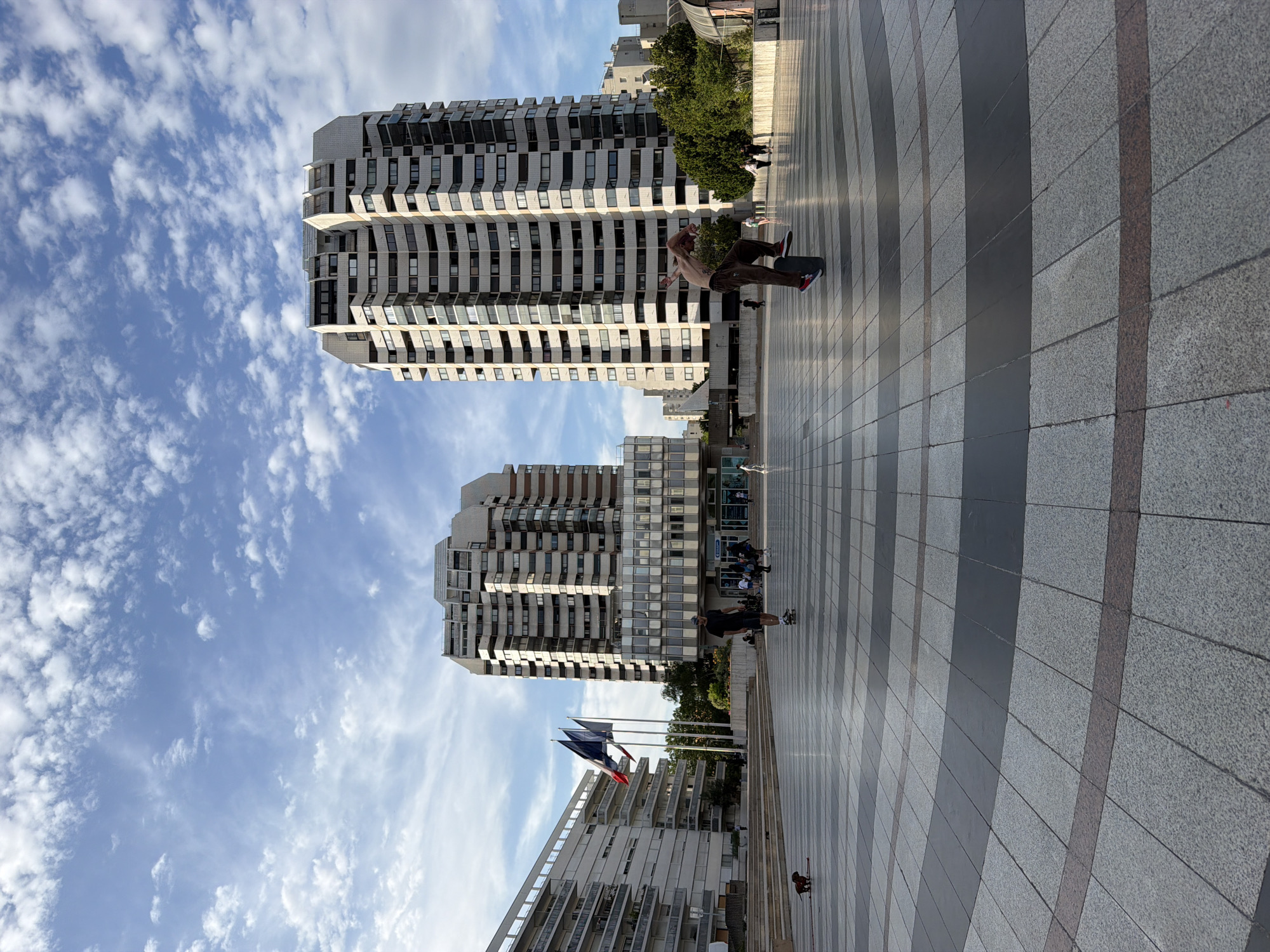 This image depicts a lively outdoor scene in Créteil, France, with a focus on modern architecture and recreational activity. Two large, multi-story buildings with distinct, undulating facades dominate the background. These structures, likely residential or office buildings, are characterized by rows of balconies and numerous windows, suggesting a contemporary urban environment.
In the foreground and midground, a paved plaza with a geometric pattern of light grey, dark grey, and reddish-brown tiles stretches into the distance. The sunlight casts long shadows, indicating it is likely late afternoon or early evening. The sky is a bright blue, dotted with fluffy white clouds, suggesting pleasant weather.
The primary activity captured is skateboarding. A person is seen mid-trick on a skateboard in the midground, appearing athletic and engaged. Further in the background, other individuals can be seen on skateboards or walking, suggesting a popular spot for recreation. Two French flags are prominently displayed on flagpoles, adding a national touch to the location. The overall mood appears energetic and social, with people enjoying the open space and engaging in leisure activities against the backdrop of impressive architecture.