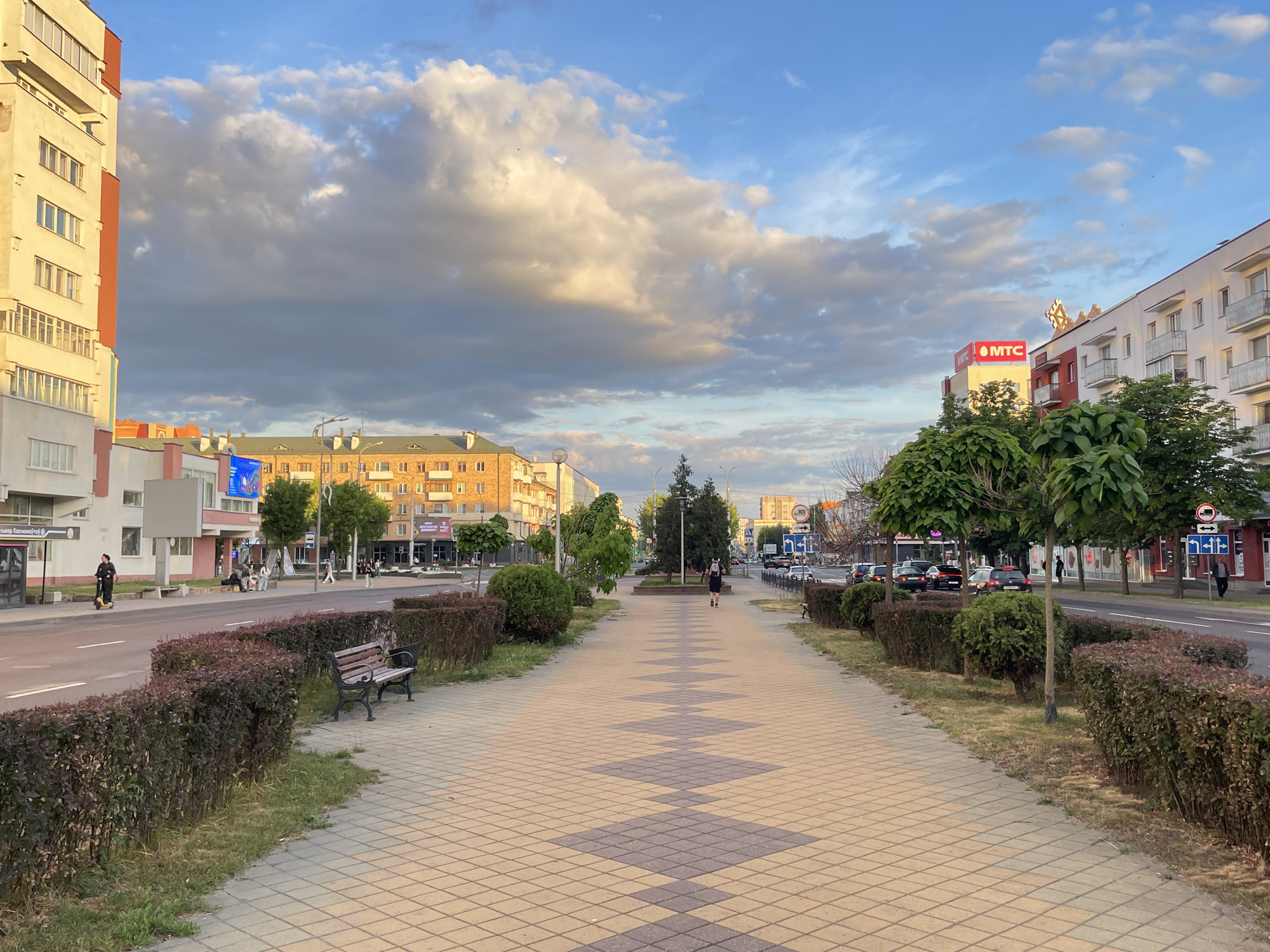 This image depicts a wide-angle street scene in Brest, Belarus, specifically on Vulitsa Kasmanaŭtaŭ (Cosmonauts Street), identified by Cyrillic signage at a bus stop. The setting appears to be late afternoon or early evening, characterized by warm, golden light illuminating the upper portions of clouds and buildings. In the immediate foreground, a broad paved sidewalk extends centrally into the distance, patterned with alternating light yellow and dark gray square tiles forming a diamond motif. To the left of the sidewalk, a dark-framed wooden park bench is positioned behind a strip of grass. This grass separates the sidewalk from a row of low, neatly trimmed hedges with dark green and reddish foliage, bordering a two-lane asphalt road. White solid and dashed lines mark the road lanes. A similar arrangement of hedges and grass borders the road on the right side. The midground features various elements. On the left side of the road, a modern bus stop shelter with a light gray roof displays "Вуліца Касманаўтаў" (Vulitsa Kasmanaŭtaŭ) in Cyrillic script. A person in dark clothing rides a yellow electric scooter on the sidewalk near the bus stop. Further down the left side, a group of approximately four individuals is seated on a bench, and a large blue billboard is visible above a building. On the right side of the road, a person in dark attire walks away from the camera on the patterned sidewalk. Several other pedestrians are visible in the distance on both sides of the street. Vehicles, including cars, are parked or moving on the road. Architectural features include multi-story buildings lining both sides of the street. On the far left, a tall building with light-colored facades and prominent orange-red vertical accents features numerous windows and balconies. Further left, other buildings exhibit yellowish facades with green pitched roofs, and some lower levels are painted light pinkish-white. On the right side of the street, multi-story buildings are predominantly white with red accents and balconies. A large red "MTC" sign is conspicuously mounted on the rooftop of a building on the right. Streetlights, trees with varying canopy shapes, and directional road signs are present throughout the scene. The background shows additional buildings receding into the distance. The sky above is a mixture of bright blue patches and large, textured cumulus clouds. These clouds display warm, golden illumination on their upper surfaces from the low sun, while their undersides appear in darker