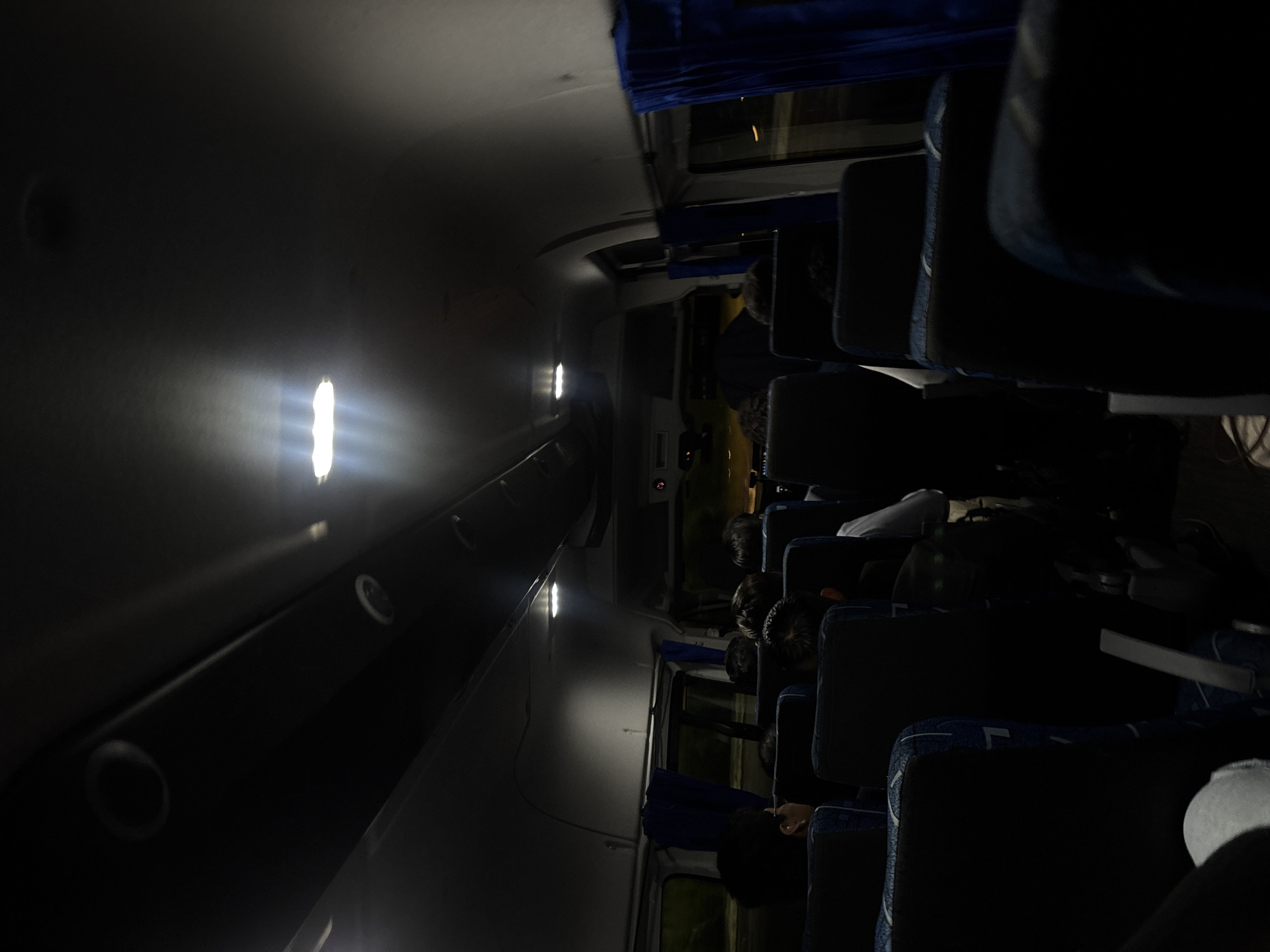 The image captures the interior of a bus or coach, taken from a slightly elevated vantage point and rotated 90 degrees clockwise, making the ceiling appear on the left and the floor on the right. The setting is predominantly dark, indicating nighttime travel.
Bright, white, linear strip lights are affixed along the upper wall/ceiling of the bus, illuminating the immediate area with a strong glow. Below these lights, a dark, elongated luggage rack extends down the cabin. The main seating area on the right side of the aisle is significantly darker, creating deep shadows.
Several passengers are seated in rows of dark, possibly blue, upholstered seats. Most individuals are visible from behind or as partial figures, their heads and shoulders discernible, suggesting they are resting or simply seated during the journey. Blue curtains are drawn on some windows next to the seats. The windows reflect only darkness, confirming the time of day. The overall scene conveys a quiet and subdued atmosphere, characteristic of overnight bus travel. There is no visible text in the image.