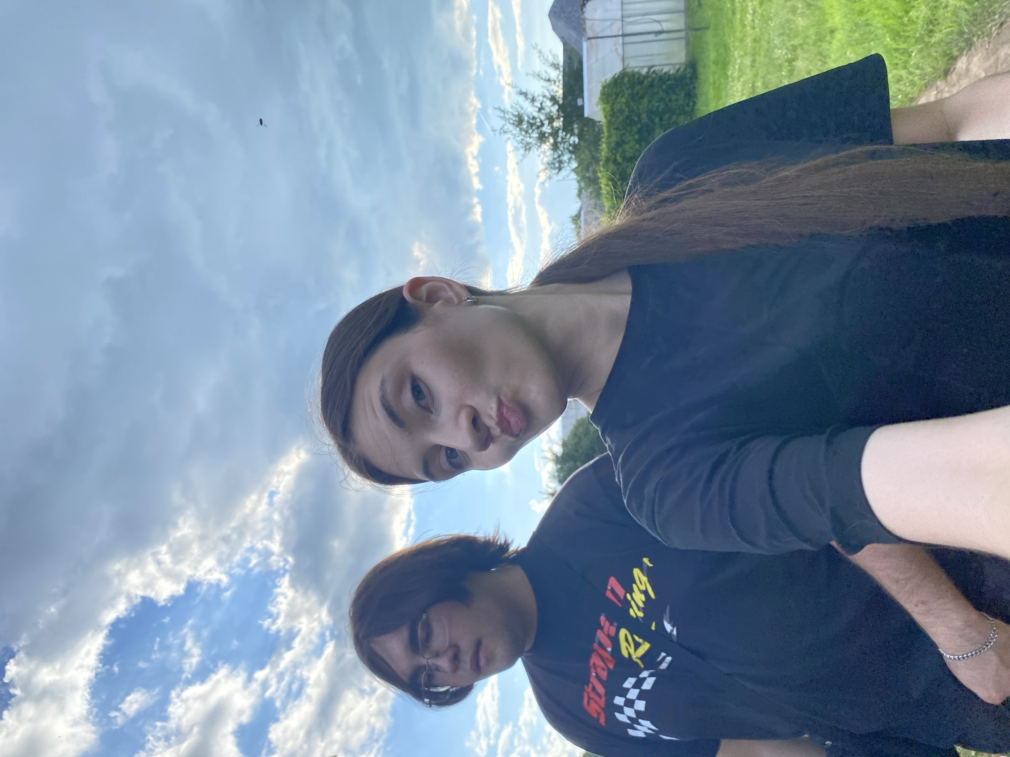 Two people are posing for a selfie outdoors. The person in the foreground on the right has long brown hair and is wearing a black t-shirt, making a kissing face at the camera. Behind them and slightly to the left, another person with brown hair and glasses is visible, also wearing a black t-shirt with yellow and red text that appears to say "Strange Racing". They are both looking towards the camera. The background consists of a blue sky with scattered white clouds and greenery, suggesting a daytime setting. A small insect is visible in the sky above the two individuals. The scene appears to be a casual outdoor gathering in Novyi Ural, Russia, possibly during the daytime with fair weather.