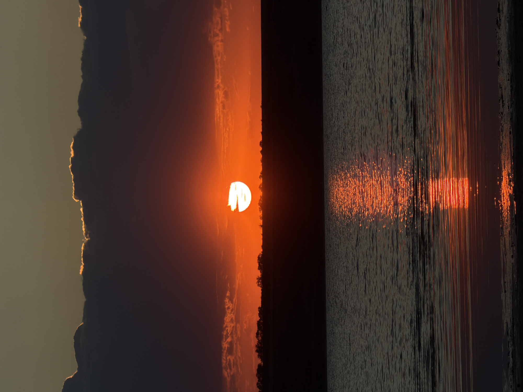 A vertical shot captures a sunset over a body of water, likely in Odesa, Ukraine. The sun, a vibrant orange sphere, is partially obscured by a dark, horizontal cloud bank, its lower edge appearing as a glowing crescent. The sky above the clouds transitions from a muted yellow to a deep orange, reflecting the sun's intensity. Below the cloud line, a dark silhouette of trees lines a distant horizon. The foreground is dominated by calm, dark water, mirroring the fiery hues of the sunset in a shimmering reflection that stretches across the surface. The overall atmosphere is serene and colorful.