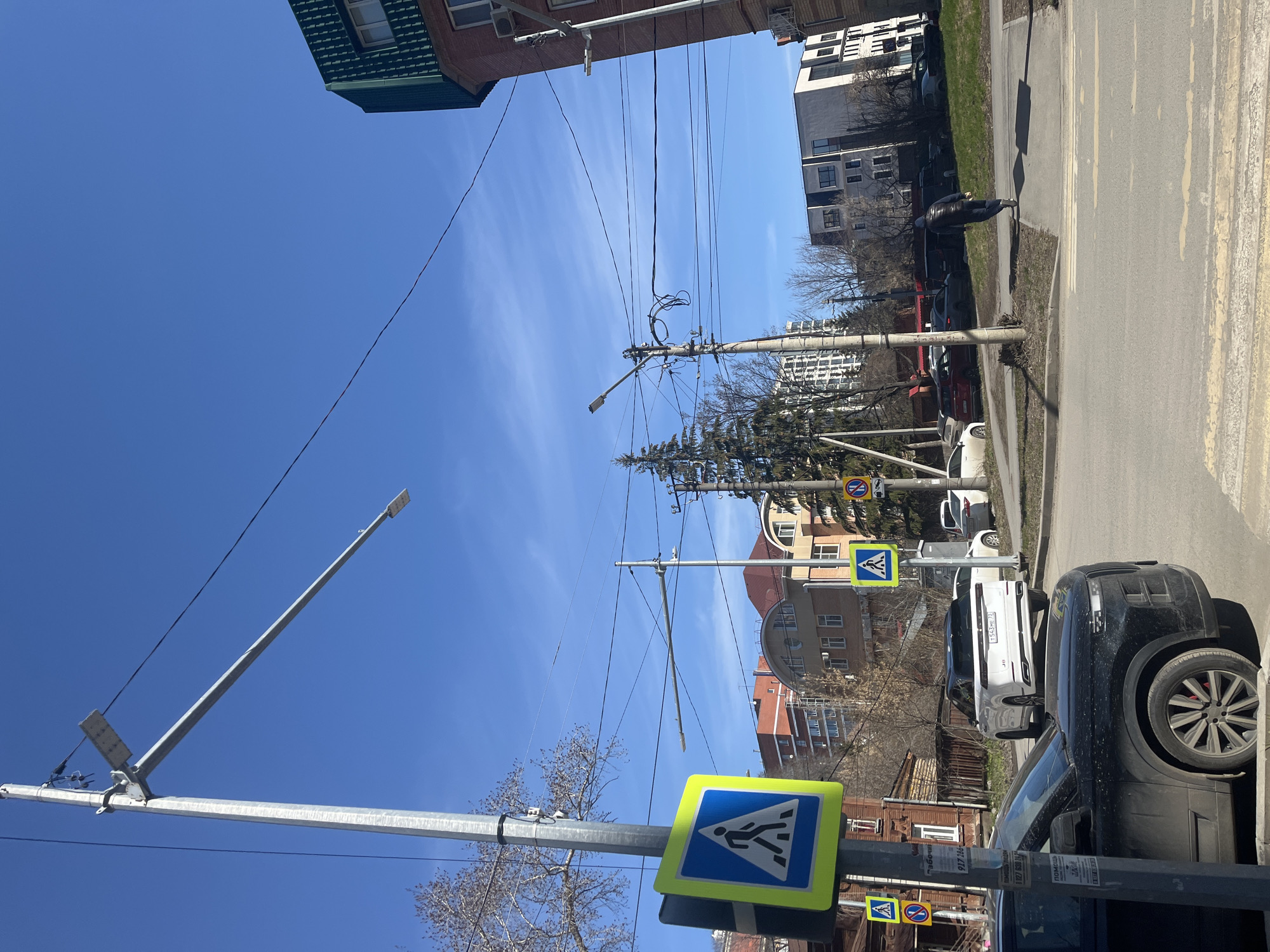 A street scene in Ulyanovsk, Russia, shows a bright blue sky above buildings, utility poles with wires, and street signs. The street has parked cars on the right side. A person walks along the sidewalk on the right. Pedestrian crossing signs are visible on poles. Buildings in the background include residential and commercial structures.