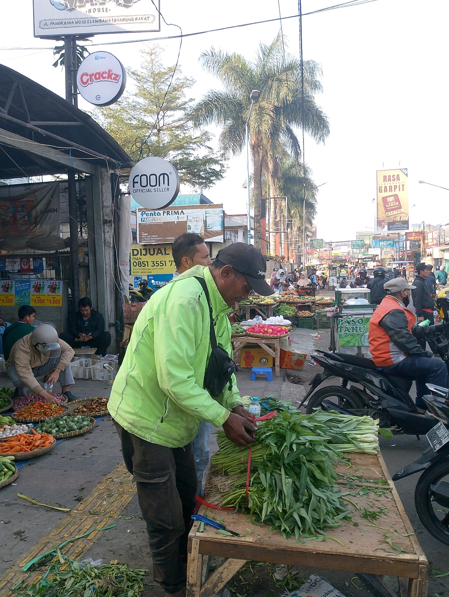 The image depicts a bustling outdoor market scene in Lembang, Indonesia, likely on a sunny day with a clear sky. The foreground is dominated by a man wearing a bright green jacket and dark pants, who is actively arranging a large bunch of leafy green vegetables, possibly water spinach, on a wooden table. His hands are visible, tied with red string, suggesting he is preparing the produce for sale. Beside him, on the ground, are scattered vegetable trimmings and a plastic water bottle.
To the left, another person wearing a light-colored top and a helmet is sorting through piles of colorful vegetables, including red onions, carrots, and possibly chilies, laid out in woven baskets on the ground. A third person in dark clothing is seated behind him, seemingly attending to the goods.
In the mid-ground, the market extends into a street lined with stalls and vendors. Several people are milling about, some appearing to be customers and others vendors. Various produce, fruits, and other goods are displayed on tables and in crates. Signs in Indonesian are visible throughout, including "FOOM OFFICIAL SELLER" and advertisements for "RASA GARPIT KRETEK." Palm trees are scattered throughout the scene, indicating a tropical or subtropical climate.
In the background, more market stalls and buildings line the street, suggesting a dense urban or semi-urban environment. Motorcycles are parked and in use, common in Indonesian urban settings. The overall impression is one of lively commerce and daily life in a Indonesian market.