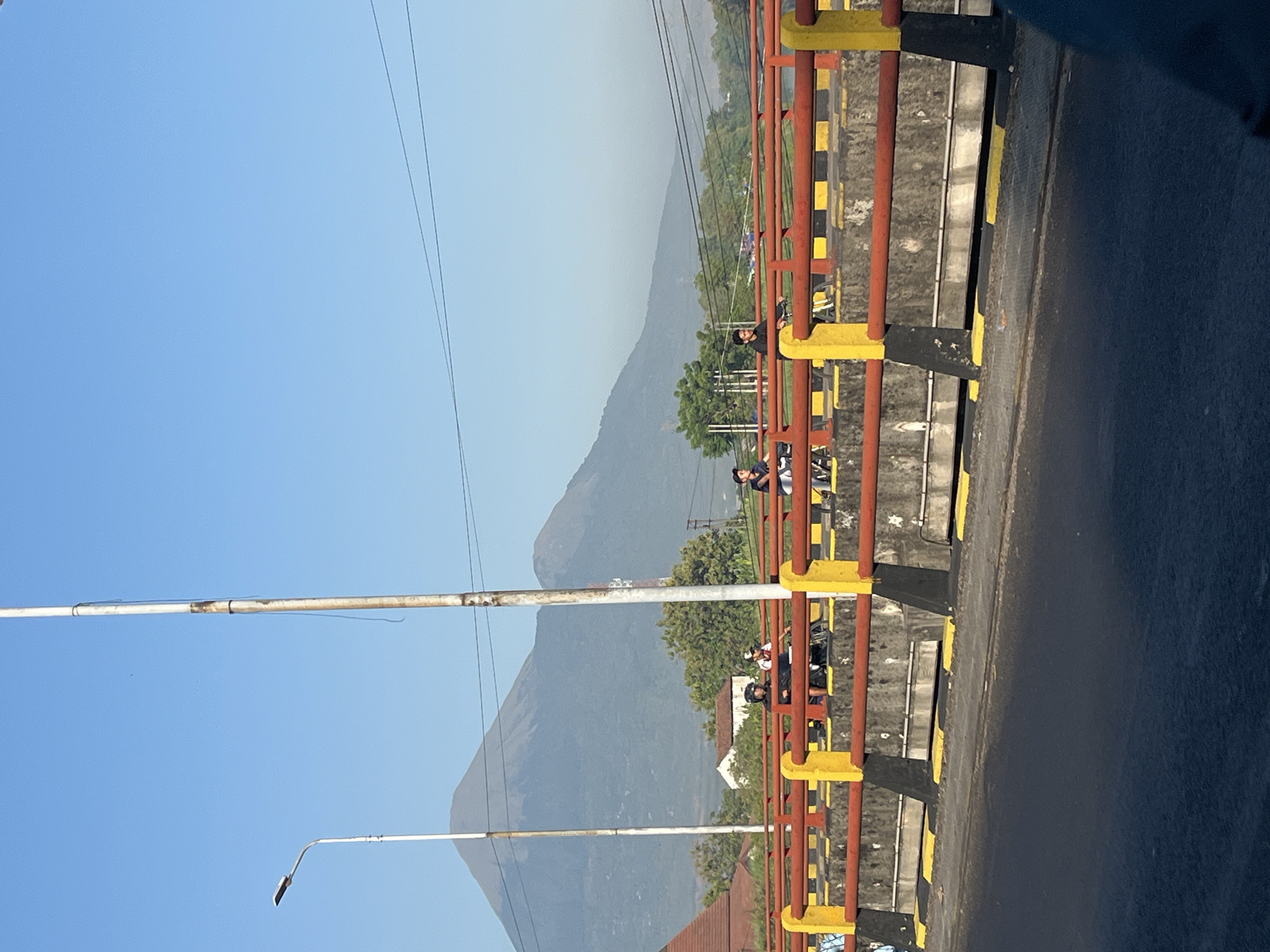 In Gempol, Indonesia, a bridge features people on bicycles. The bridge has a red and yellow railing with black and yellow striped markings along its base. Behind the bridge, a mountain range is visible under a clear blue sky. A street light stands on the left side of the image, and a tall pole, possibly a utility pole, is positioned in the center. The road surface of the bridge is dark asphalt, with some white markings. The overall scene suggests an outdoor activity on a bridge with a scenic mountainous backdrop.