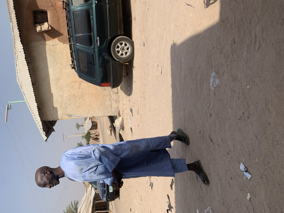 The scene captures an unpaved, dusty street in Bojude, Nigeria, under clear daytime conditions. A bald, dark-skinned adult male, wearing a light blue long-sleeved tunic over matching trousers and dark green flip-flops, walks from right to left, smiling and looking towards the left off-frame. His hands are clasped at the waist. Behind him, a dark green station wagon with a roof rack is parked, facing right, adjacent to a single-story, earth-toned building with a corrugated metal roof and a small, open window with wooden shutters. A light green pole topped with a solar street light extends upwards above this building. In the background, a dark sedan is partially visible on the unpaved road, near other simple structures with corrugated roofs and several palm trees. The ground is scattered with miscellaneous debris including plastic and paper, and distinct shadows are cast from the right side of the frame.