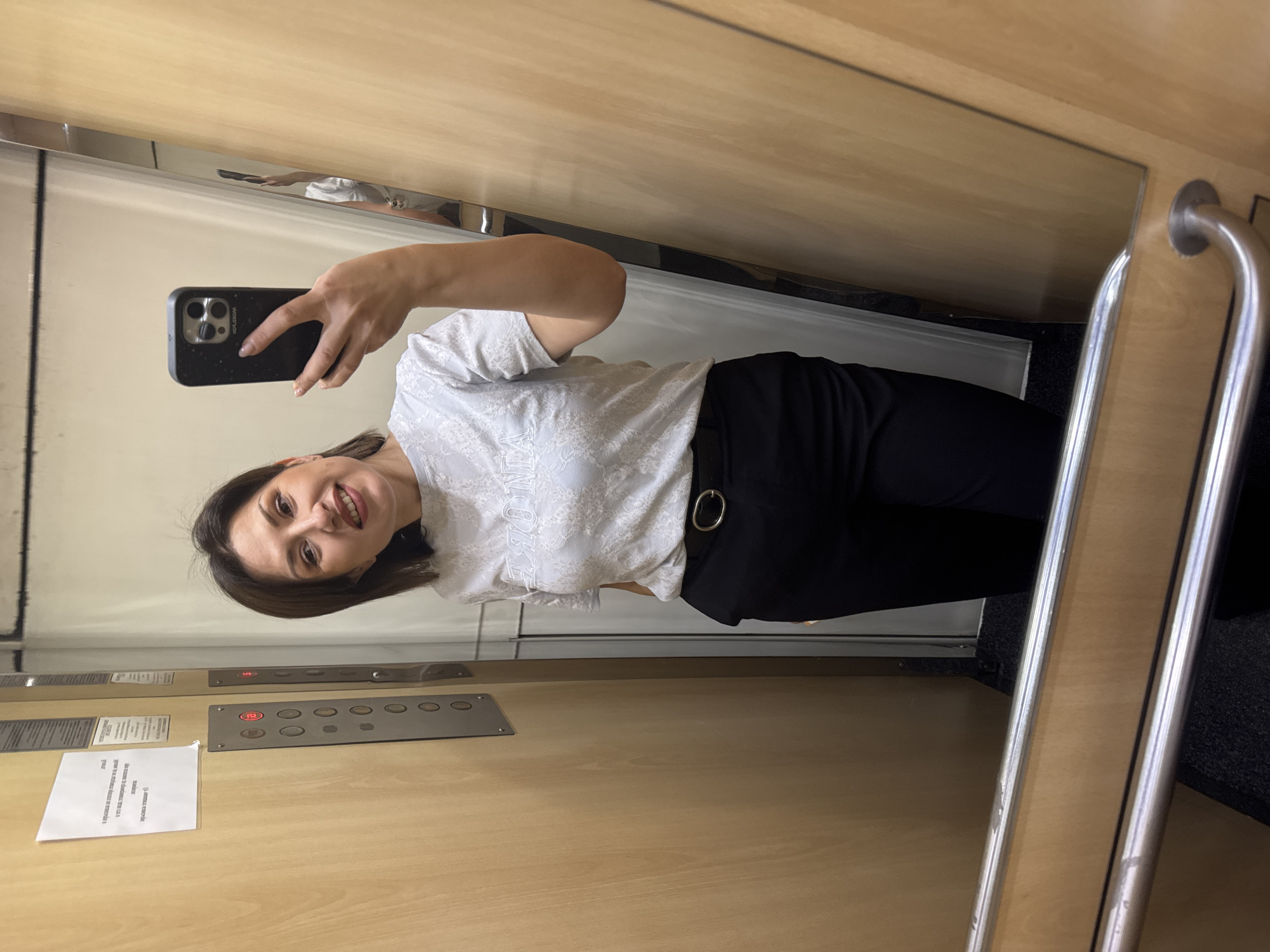 A woman is taking a selfie in an elevator. She is wearing a light-colored patterned top and black pants with a circular belt buckle. She is holding a black smartphone in her right hand. The elevator has wood paneling, a mirrored wall, and a stainless steel grab bar. A control panel with buttons is visible on the wall. The background suggests the inside of an elevator, and there are no specific indicators to pinpoint the exact location in Sofia, Bulgaria.