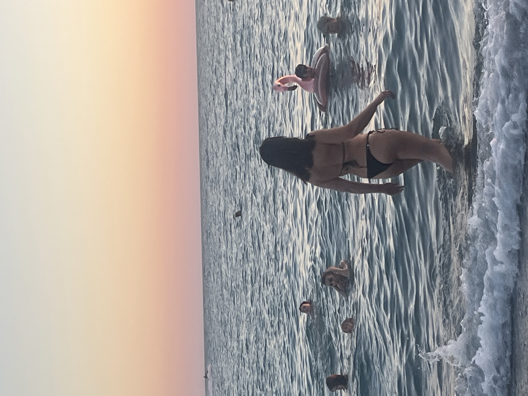 A wide shot captures a beach scene in Sochi, Russia, at sunset. The sky is a gradient of pale yellow to soft pink, meeting the calm, blue ocean. Several people are visible in the water. In the foreground, a woman in a black bikini walks towards the camera, wading through shallow waves. In the middle ground, a pink flamingo float carries a person, and another person wearing a cap is nearby. Further back, more individuals are swimming or floating, with a jet ski visible in the distance on the left. White waves break at the bottom edge of the frame.