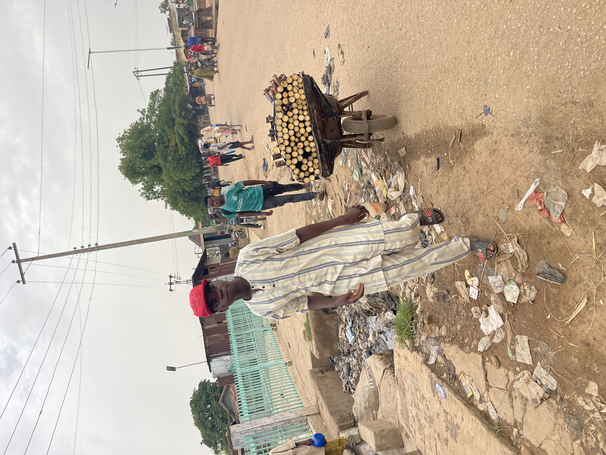The image depicts a street scene in Chikaji, Nigeria. The foreground features a man in a striped shirt, matching pants, and a red baseball cap. He is standing next to a sidewalk littered with trash. Behind him and to the right is a wheelbarrow loaded with sugar cane stalks. The background shows a dirt road with people walking. There are buildings, trees, and power lines. The sky is overcast.