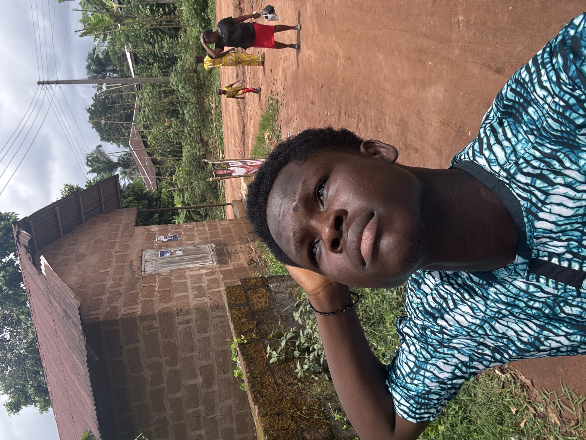 A selfie taken by a dark-skinned young man. He is wearing a blue and white patterned shirt. In the background, there is a dirt road with three people walking away from the camera. A small brick building is on the left. The location appears to be in Eziudo, Nigeria.