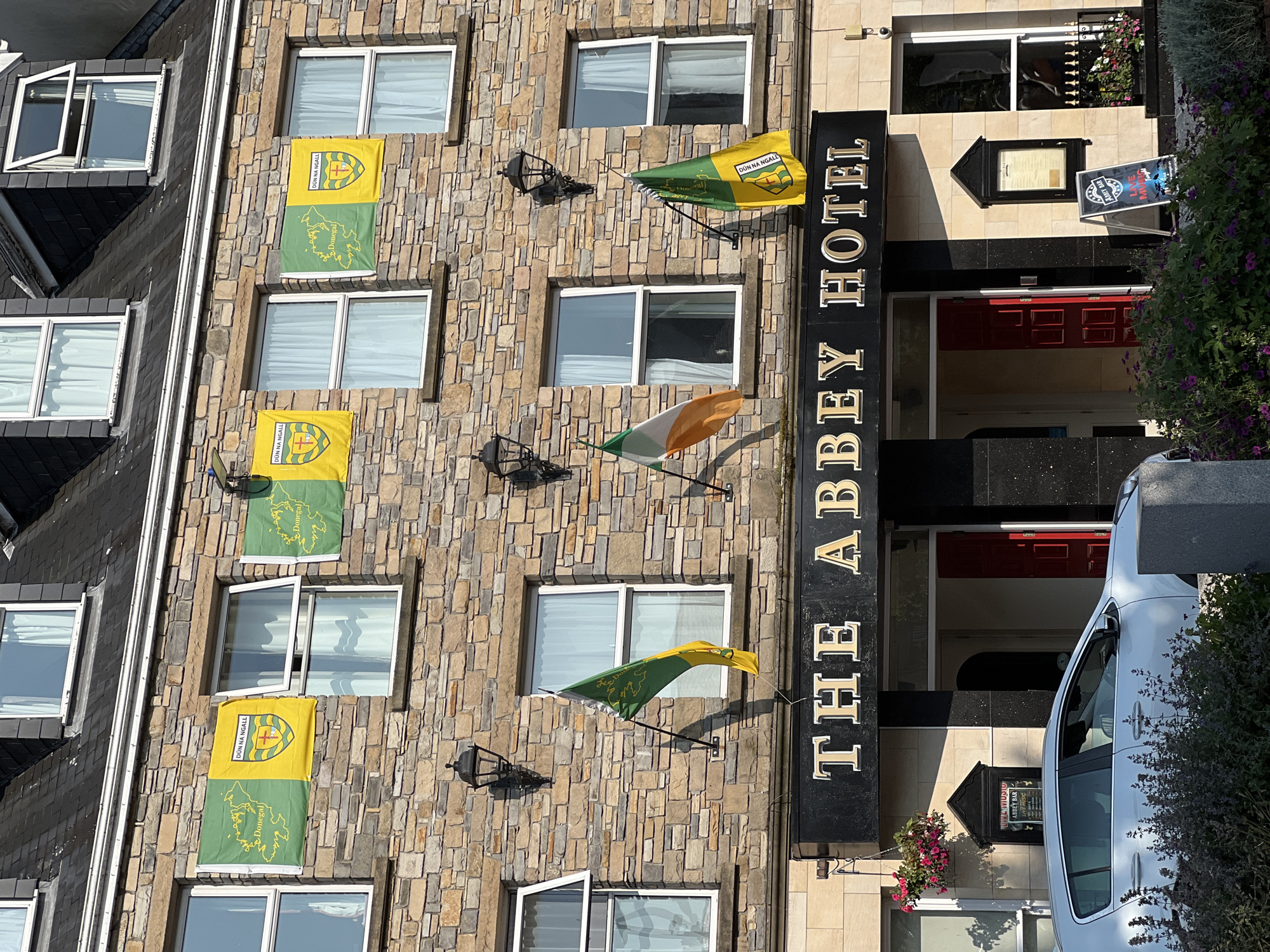 The image shows the facade of "The Abbey Hotel" located in Donegal town, Ireland. The hotel is a multi-story building constructed with a stone facade. Several flags are displayed on the exterior walls, including flags of Donegal County and the Irish national flag. The hotel entrance features red double doors and signage. A white car is parked in front of the hotel, and there are flower planters and vegetation surrounding the entrance. The scene is bathed in sunlight, casting shadows on the building and the ground.