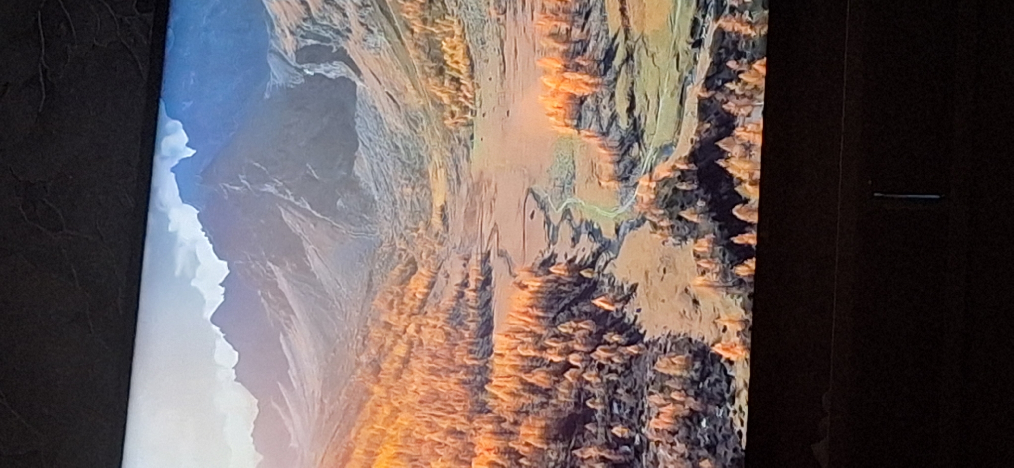 The image displays a scenic mountain valley with autumn foliage. The foreground features a dense cluster of trees with vibrant orange and yellow leaves, illuminated by warm sunlight. A winding dirt path or stream meanders through the valley floor, bordered by patches of dry grass and scattered trees. In the background, majestic, rocky mountains rise towards a sky with soft, hazy clouds. The overall impression is one of natural beauty during the fall season.
There is no discernible connection to the city of Bila Tserkva, Ukraine in this image. The scene is a landscape, likely a national park or wilderness area, and lacks any urban elements or identifying landmarks specific to that location.