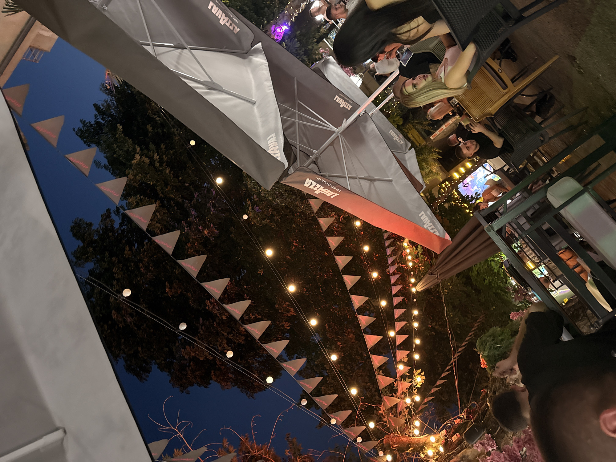 The image depicts an outdoor seating area, possibly a cafe or restaurant, at night. The scene is decorated with strings of lights and triangular flags, creating a festive ambiance. Several tables and chairs are visible, with patrons seated at some of them. Two individuals, a man and a woman, are the most prominent figures in the foreground, looking towards the right side of the frame. Umbrellas, some bearing the "Lavazza" logo, are also present, providing shade. The background features trees and the night sky. The location is identified as Lovech, Bulgaria.