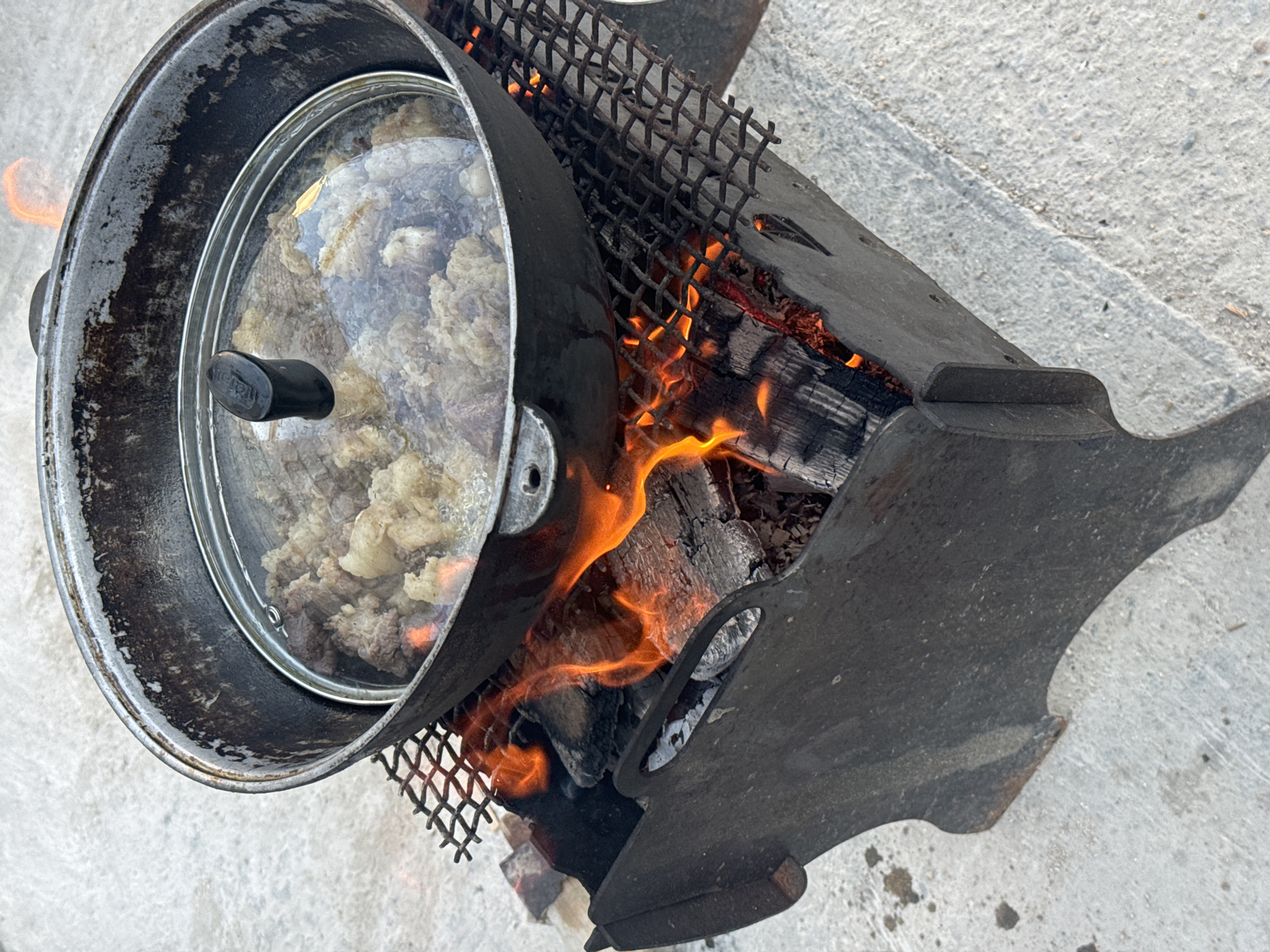 A black pot with a glass lid sits atop a metal grill over a fire pit filled with burning wood. The pot contains chunks of meat, which appear to be cooking. The scene is outdoors, with a concrete surface surrounding the fire pit. The overall impression is one of outdoor cooking. There are no people visible in the image.