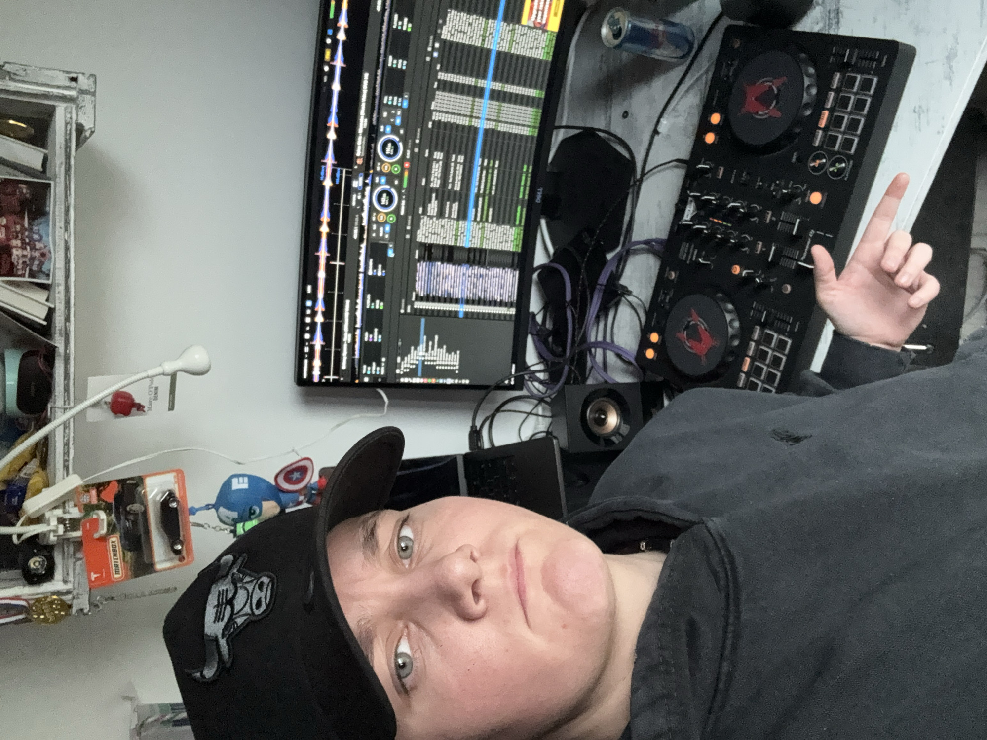 A person wearing a black baseball cap and a black hoodie is taking a selfie with a DJ controller, computer monitor, and other electronics visible in the background. The person is oriented at an angle in the frame with their face toward the bottom left and their hand raised toward the upper right. The monitor displays data lines and symbols. A gray wall is behind the monitor and controller. A small shelf with miscellaneous objects is to the left of the monitor. The setting of this image cannot be reliably discerned, therefore it is not possible to describe the location as being in the city of Sofia, Bulgaria.