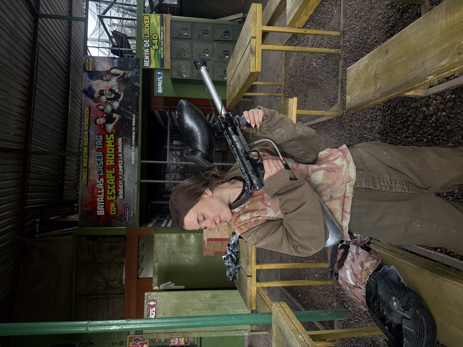 A woman in a brown jacket and pants holds a paintball gun, resting on a wooden picnic table. Behind her, a sign advertises "Laser Tag" and "Escape Rooms." Additional text on the sign reads "Atrévete y cumple la misión" and encourages purchasing tickets. A sign advertises locker rentals for $40. A camo backpack and black helmet rest on the bench next to the woman. The area appears to be outdoors with gravel ground.