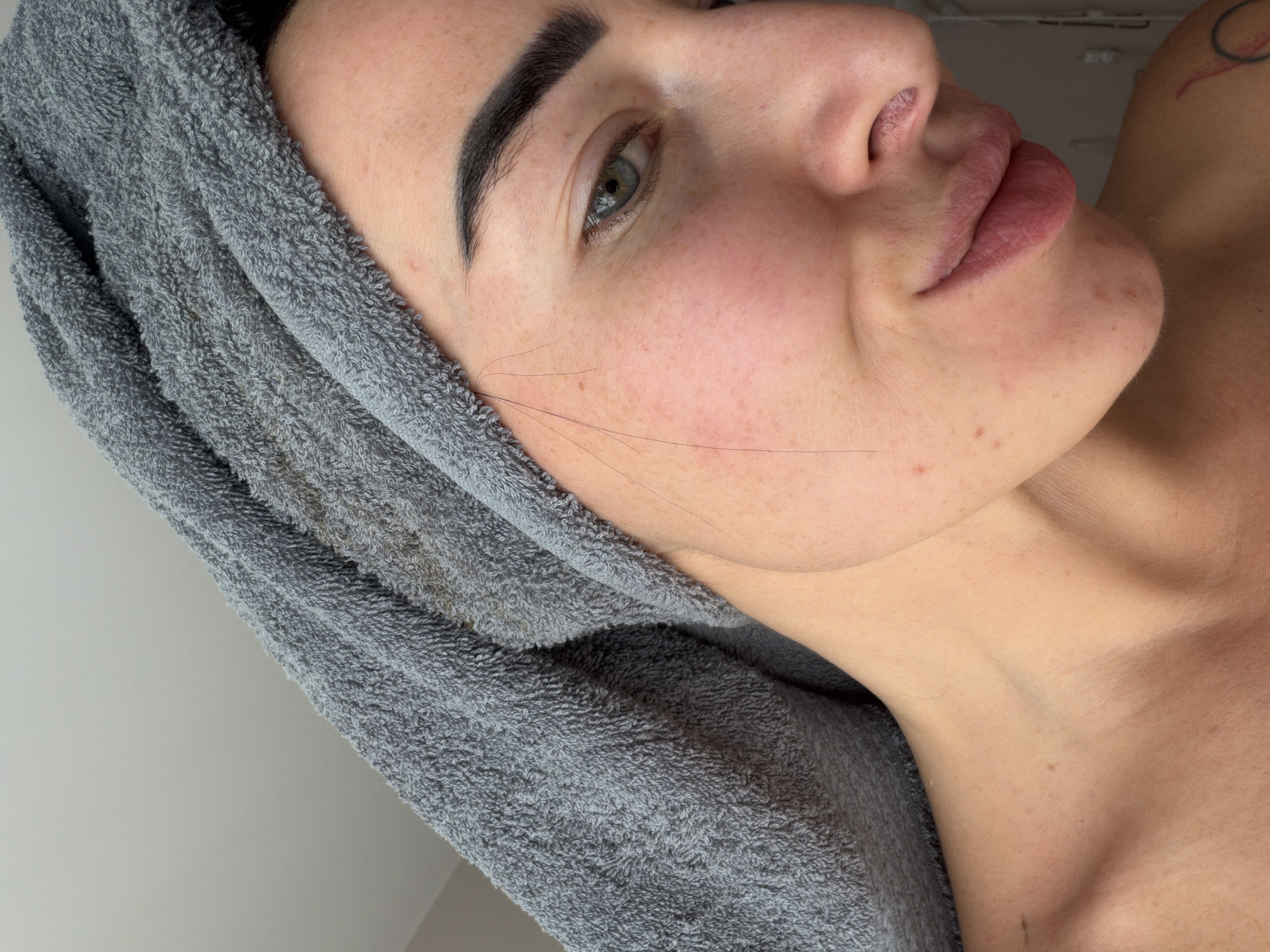 This media file, originating from an unknown location in Russia, is a close-up, tilted portrait of a person, likely a woman, presented in an almost vertical orientation. The subject's head is wrapped in a soft, grey terry cloth towel, suggesting she may have just showered or undergone a beauty treatment. Her left eye, a striking green, is open and directed towards the viewer, framed by a dark, well-defined eyebrow. Her fair skin is visible, adorned with scattered freckles across her nose and cheeks. Several fine, dark hairs are noticeable on her right cheek, extending towards her hairline.
Her full lips have a natural pink hue and show some texture. The lower part of her face, neck, and upper chest are visible, appearing bare. A partial view of a red and black circular tattoo is discernible on her left shoulder or upper arm. Her expression is neutral and calm. The background is out of focus, appearing as a plain, light-colored surface, indicating an indoor setting. The lighting is soft and even, consistent with natural daylight. There is no visible text in the image.