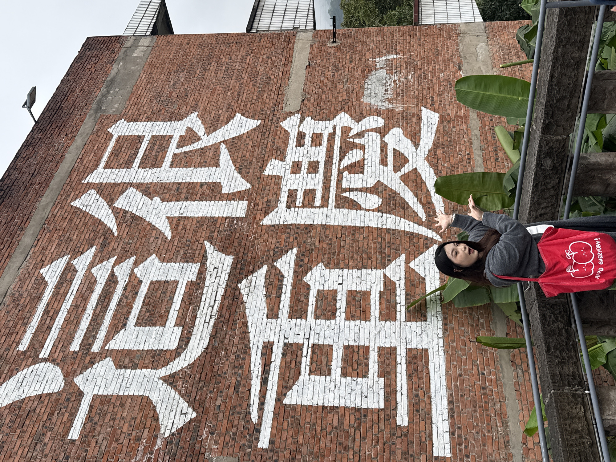 A young East Asian woman, dressed in a grey hooded cardigan over a white top and dark lower garments, poses with a red "APPLE EVERYDAY!" tote bag on her left shoulder. She stands smiling with both hands gesturing upwards, positioned behind a grey metal railing with dark stone posts. The background is dominated by a multi-story reddish-brown brick wall located in Nan'an District, China. Prominently displayed on the wall are large white Chinese characters painted vertically, reading "这 很 重 庆" (This is Chongqing). Between the railing and the wall, large green, broad-leafed plants are visible. The upper portion of the brick wall features concrete banding and a street lamp fixture on its top edge. A cantilevered metal balcony structure with a railing is visible on the far upper right side of the wall. The sky above is overcast, with distant green foliage and a building structure visible on the far right.