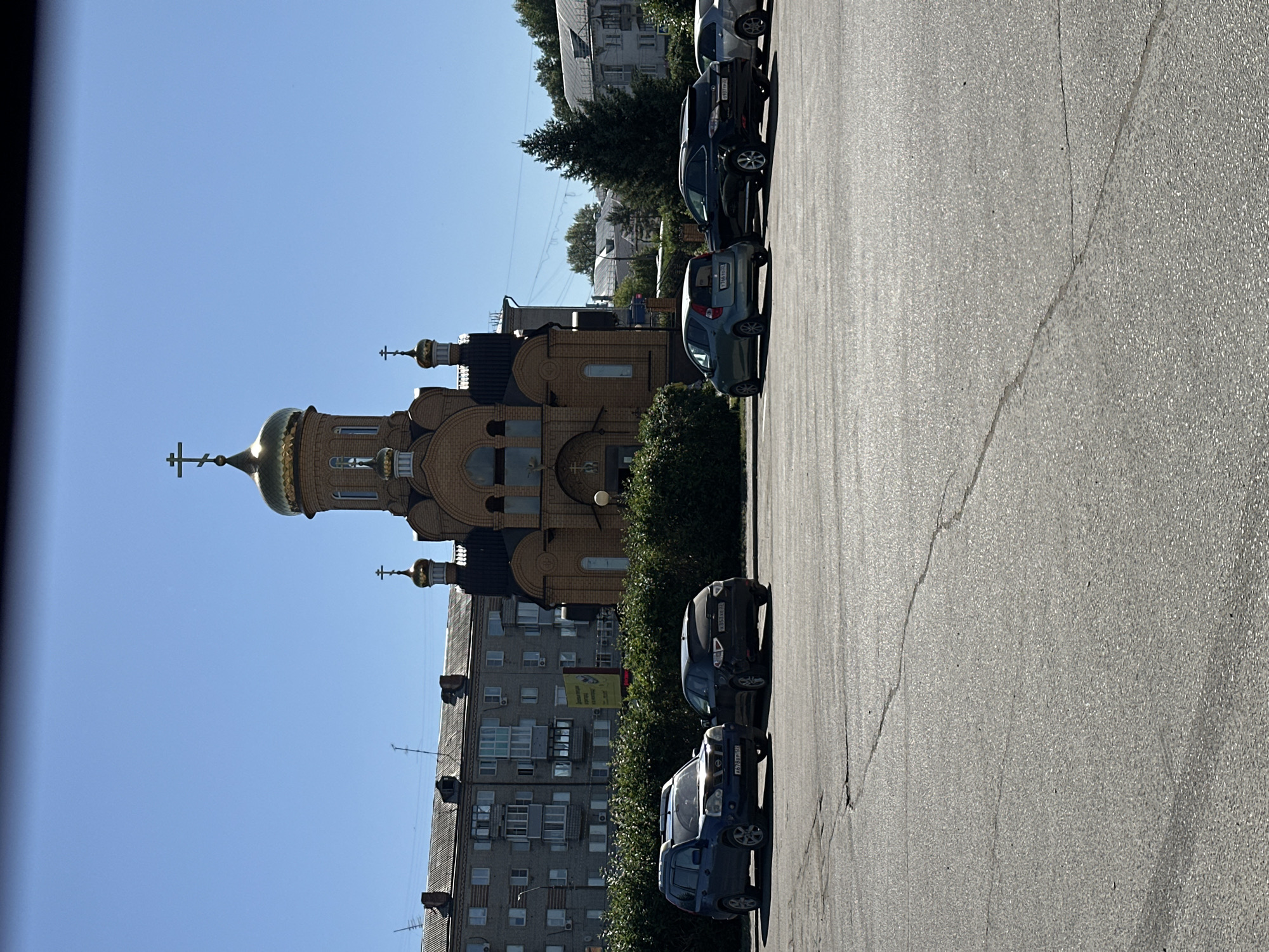 A bright, sunny day in Novoaltaysk, Russia, features a large, brick Orthodox church with a golden dome and cross as the central focus. The church is flanked by residential buildings and trees. In the foreground, several cars are parked on an asphalt lot, which shows signs of wear and cracking. The scene appears to be in a public space, likely a parking lot adjacent to the church or a nearby civic area.