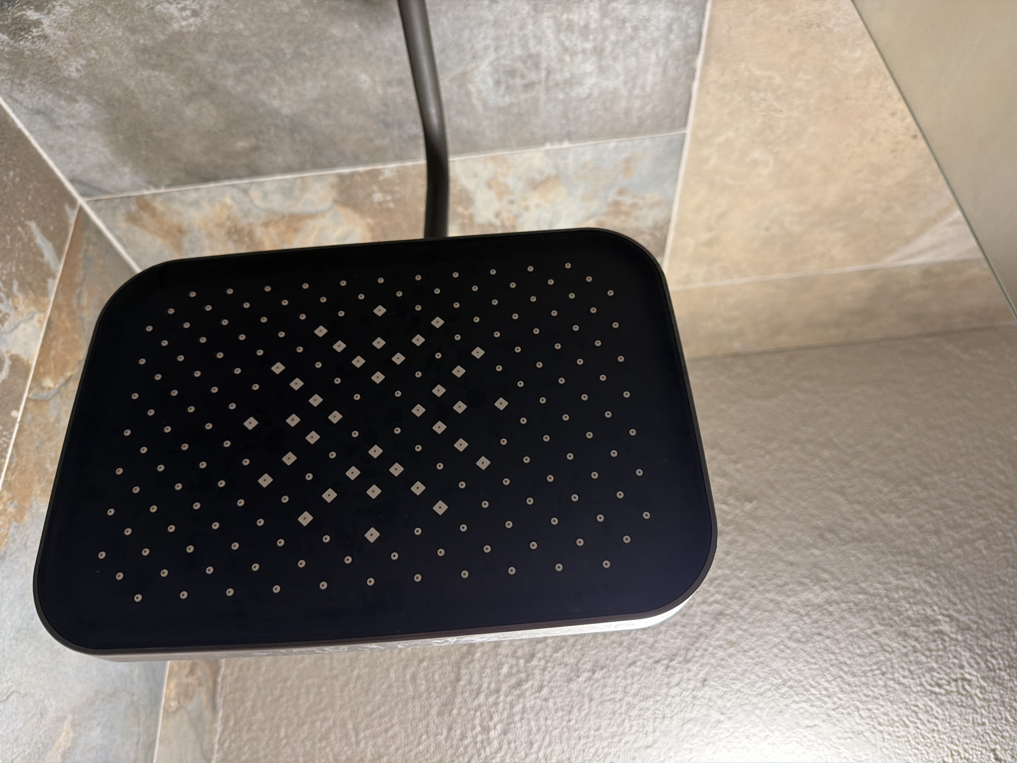 The image shows a close-up, overhead view of a modern, black rectangular shower head. The shower head has a matte finish and is dotted with numerous small, circular nozzles. Some of the nozzles are arranged in diamond-shaped patterns, while others are placed in a more scattered grid. The shower head is mounted on a dark-colored shower arm, which extends from the right side of the frame. The background consists of tiled walls, with the tiles in the upper portion of the image appearing to be a darker, slate-like gray, and the tiles in the lower portion being a lighter, beige-brown color with a textured, natural stone appearance. The lighting suggests an indoor environment, likely a bathroom. There are no people or animals present in the image. The scene is static. The specific location within the city of Sofia, Bulgaria, cannot be determined from the visual information provided.