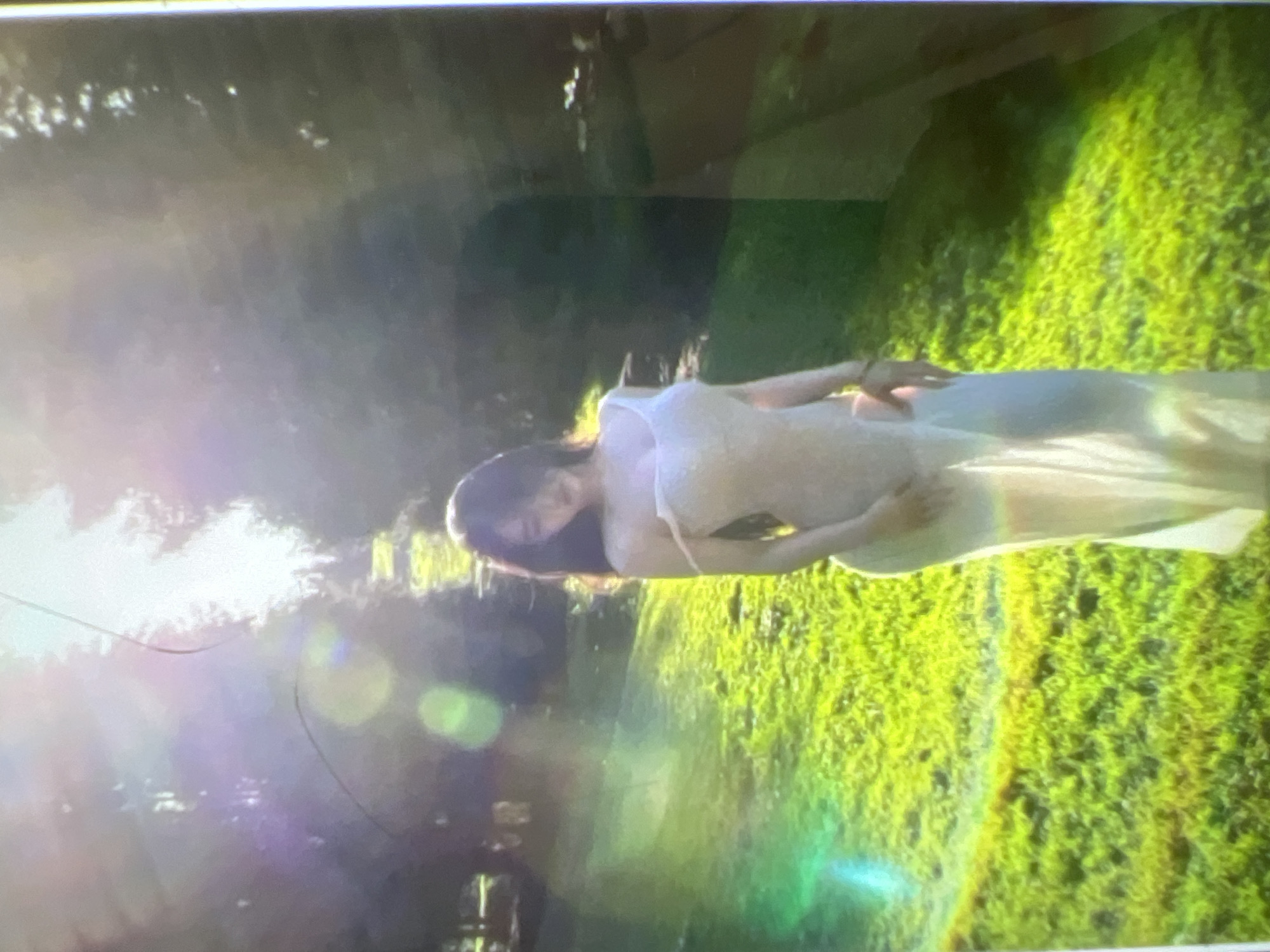 A woman in a long, sheer white dress stands in a grassy, sunlit outdoor area. The dress appears to be made of a delicate, translucent fabric, with thin straps that fall off her shoulders. Her hair is dark and cascades down her back. She is facing slightly away from the camera, with her head tilted down. The background consists of lush green grass and trees, with strong sunlight creating lens flares and bright spots throughout the image. The setting suggests a natural, possibly park-like environment. The image is undated and unlocated.
