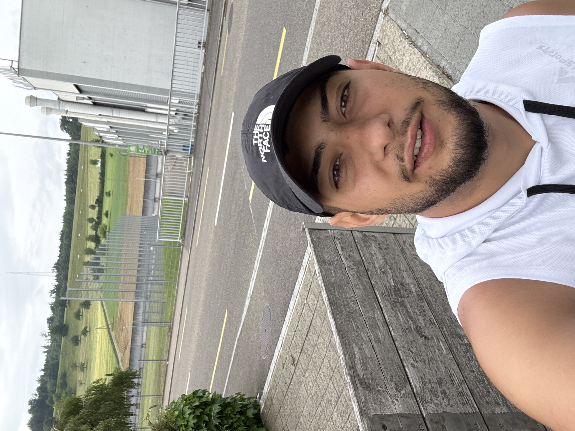 The image is a selfie of a man wearing a black "The North Face" cap and a white sports shirt. He has a beard. The background shows an asphalt road with yellow and white lane markings, a field, a building with visible pipes, and a fence. A transmission tower is visible on the horizon. The man is sitting on a wooden bench. The location is likely Lausen, Switzerland, given the landscape and the presence of the transmission tower, similar to those found in the area.