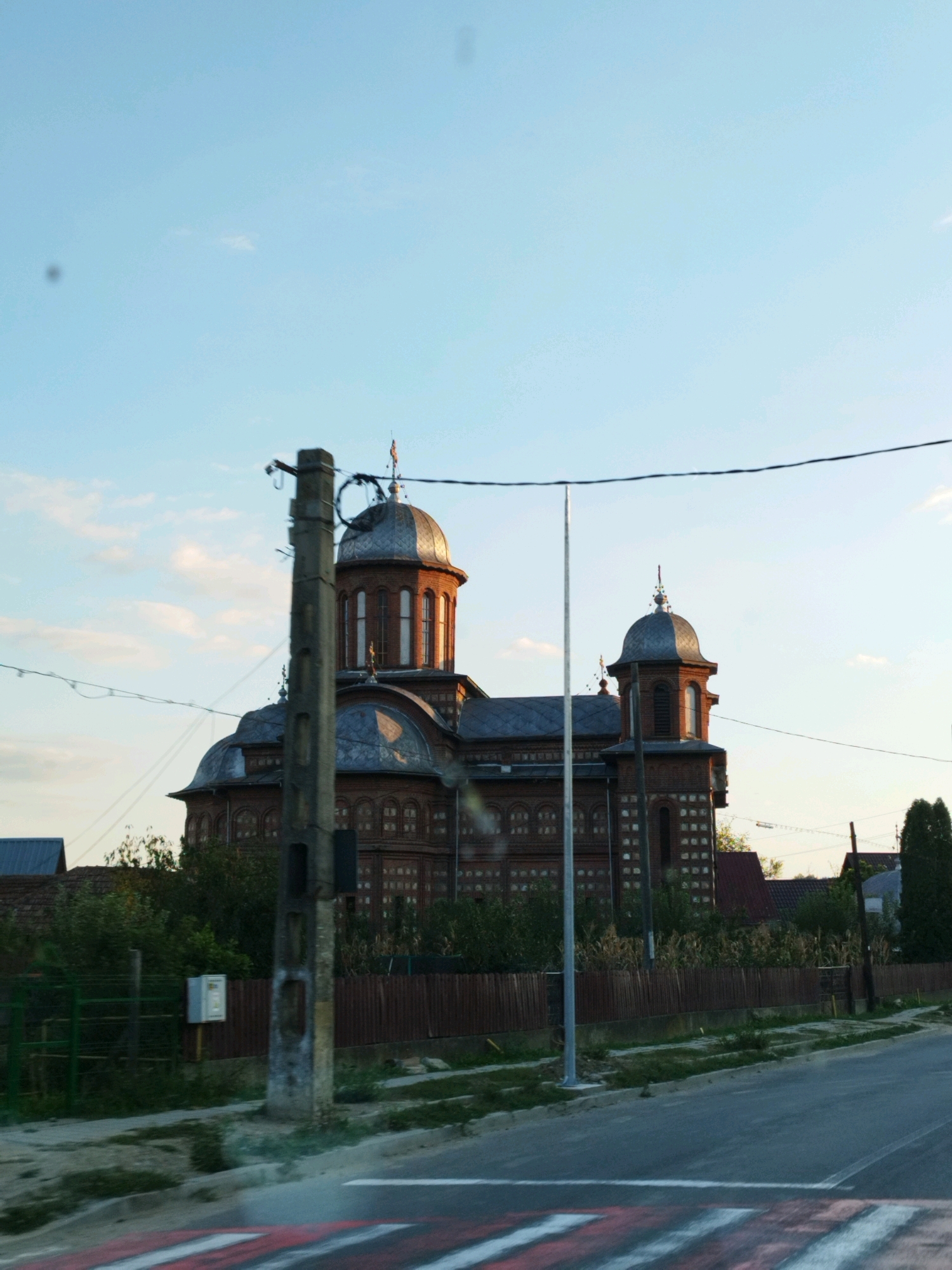 The image captures a view of a brick church with multiple domes, possibly in Curtea de Argeș, Romania, under a clear sky with scattered clouds. In the foreground, a concrete utility pole stands prominently, with wires extending across the frame. A portion of a road with a pedestrian crossing is visible at the bottom. Behind the pole, a section of the church's facade is visible, showing arched windows and decorative brickwork. A wooden fence and vegetation line the area in front of the church. The lighting suggests it is late afternoon or early evening, with warm light illuminating the church domes.