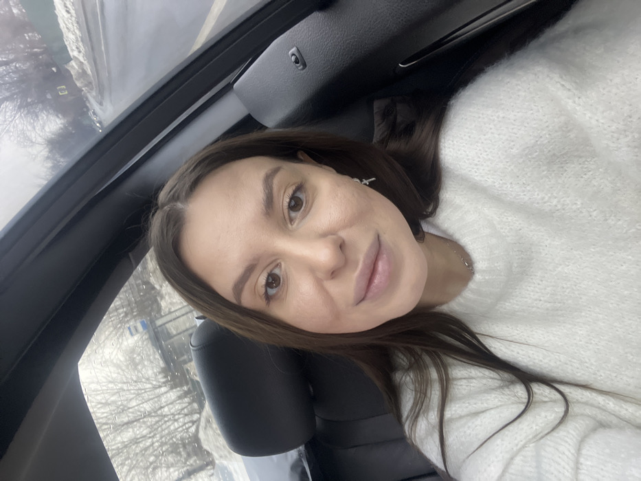 The image displays a female subject taking a selfie from within a vehicle, situated in what appears to be Vysokovo, Russia. The subject, a female with long, straight dark brown hair parted in the middle, light complexion, and brown eyes, is depicted from the upper chest up. She wears a white, chunky knit sweater, a thin silver chain necklace, and a small, silver-colored cross-shaped earring in her right ear. Her expression is neutral, with her lips slightly parted.
The interior of the vehicle shows a dark-colored seat headrest to her left and a dark-colored interior panel to her right. The background, visible through the vehicle's windows, portrays an outdoor scene during what appears to be winter or early spring. Water droplets are visible on the window glass. The environment consists of a wet asphalt road, flanked by piles of snow. Bare trees line the roadside. A blue and white bus stop sign, featuring a stylized bus icon, is partially visible through the rear window on the left side of the frame. Further along the road on the right, a green fence or wall is discernible, and distant vehicles are faintly visible. The sky is overcast and grey.