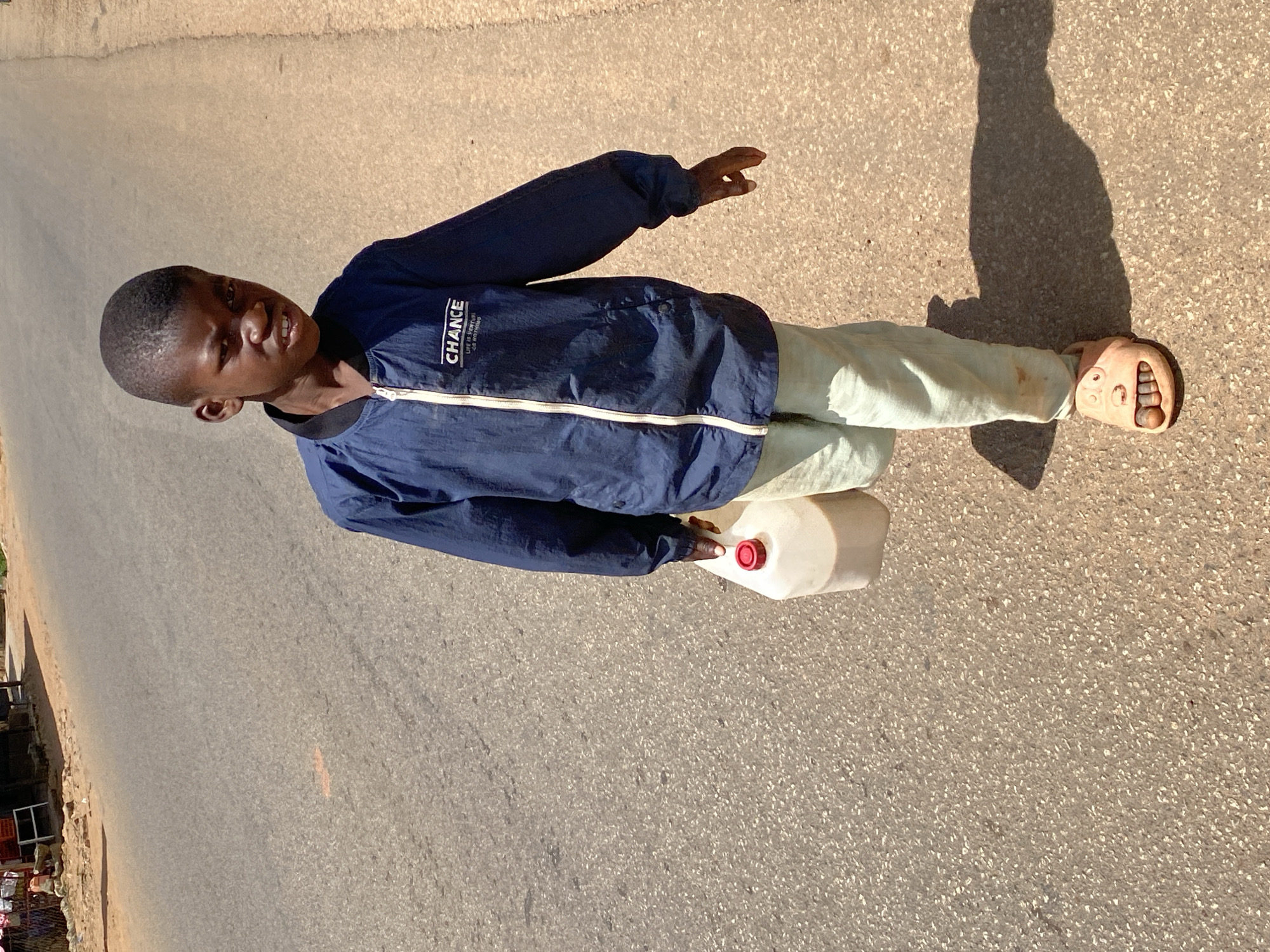A young boy, appearing to be of African descent, walks on an asphalt road in Bukuru, Nigeria, carrying a plastic jug with a red cap in his left hand. He is wearing a dark blue jacket with the word "CHANCE" and smaller text below it printed on the left breast, and light-colored pants. He is also wearing light-colored sandals that appear to have faces on them. The boy is looking directly at the camera with a grimace on his face. The road is cracked and has some debris on it. Sunlight casts a shadow of the boy and the jug on the road. In the background, there are some buildings and trees. The overall scene suggests a dry, sunny day.