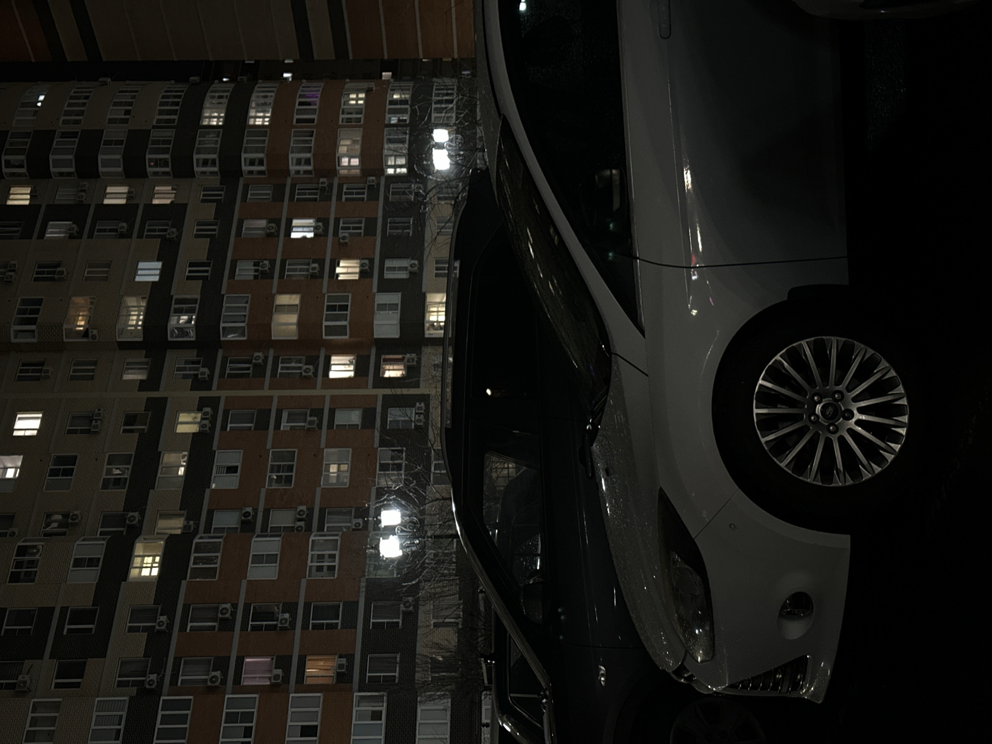 A nighttime photograph depicts a car parked in front of a multi-story apartment building, likely in Kuleshovka, Russia. The building is constructed with light-colored materials and features a pattern of windows and air conditioning units on its facade. The car, which is a light color, takes up a substantial portion of the frame. The scene is illuminated by streetlights.