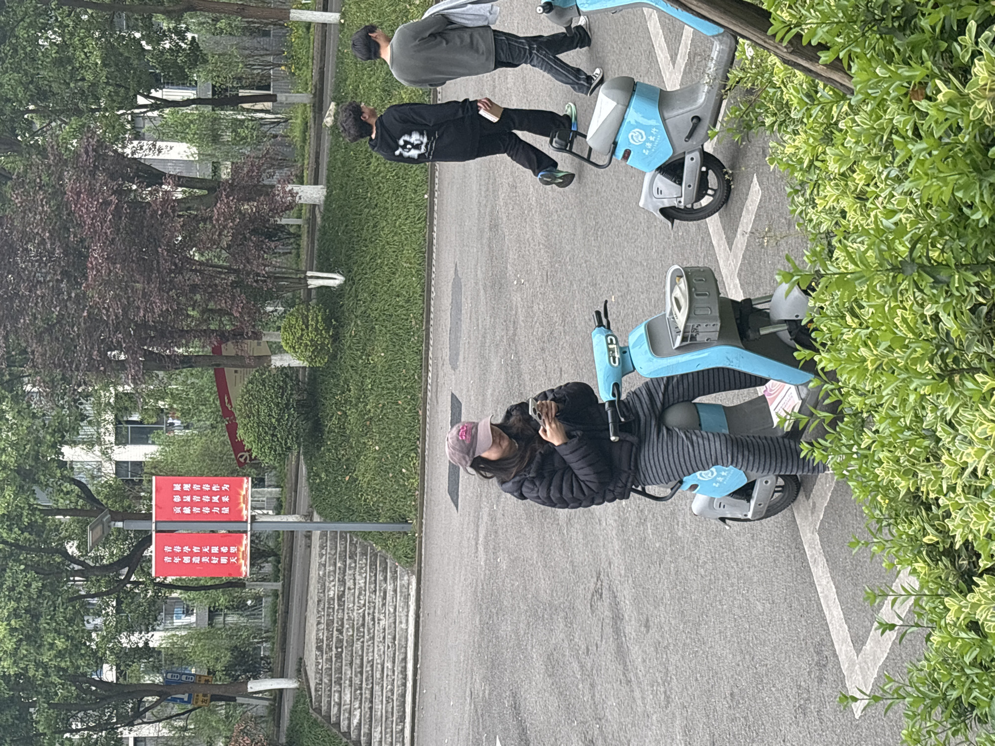 The image shows a street scene in what appears to be Jiangjin, China, featuring three people and several light blue rental e-bikes parked in designated spaces. A woman in a black jacket, striped pants, and a pink baseball cap is seated on an e-bike and looking at her phone. Two men are walking on the road alongside the parked e-bikes; one is wearing a black shirt and pants while the other is in a grey top and black pants. There is green grass and trees in the background, and a red sign written in Chinese is visible near the street.