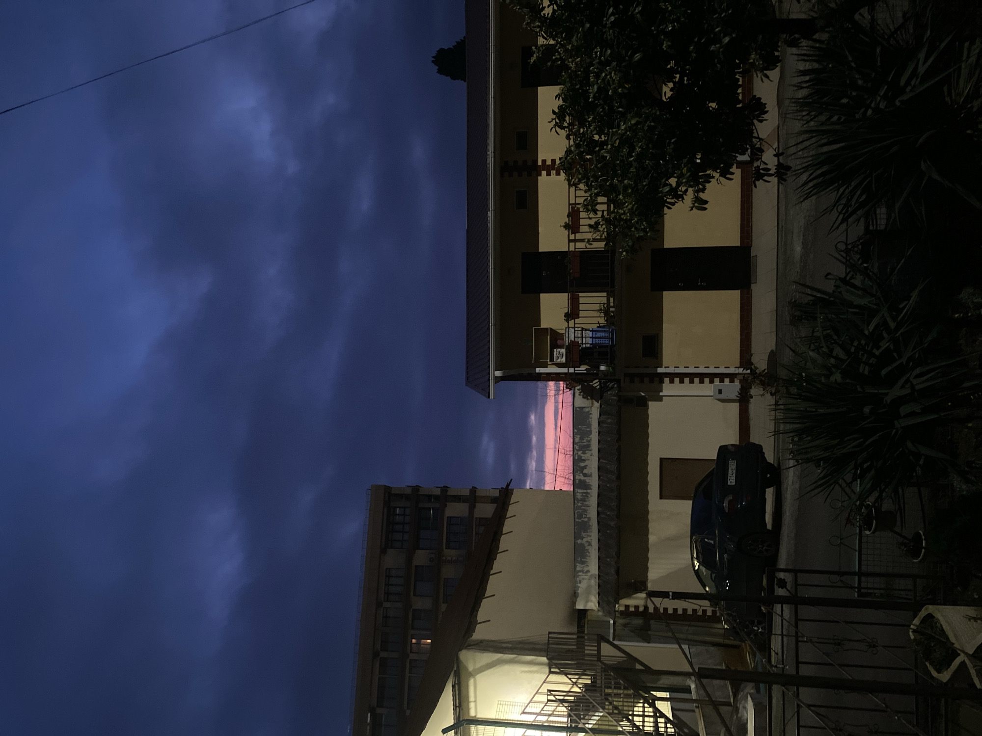 The image shows an urban scene, likely in Sochi, Russia, based on architectural styles and a license plate partially visible on a dark-colored car parked near a building. Two multi-story buildings frame a narrow view of the sky, which is dominated by a dark, cloudy dusk or night sky with a small sliver of pink or reddish glow above the horizon. The building on the right is a lighter beige color with dark window and door accents and has a small balcony on the middle level. A tree with dark foliage is partially in view near the top right of the frame. The building on the left appears to be taller, with rows of windows. A metal staircase is visible on the lower left of the frame. The scene appears to be at dusk or night.