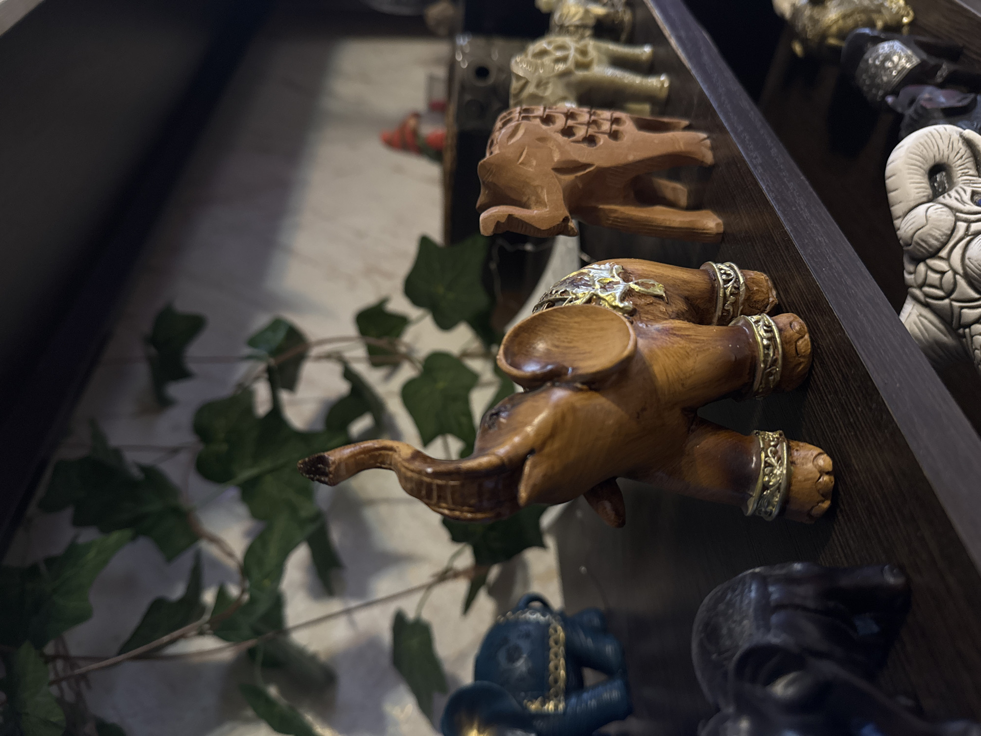 The image shows a shelf displaying various ornamental figurines, including elephants and other animals. The elephants appear to be made of wood, with gold-colored embellishments around their legs and back. Green foliage is visible behind the figurines, providing a backdrop. The scene appears to be indoors, possibly in a home or a shop. The image does not contain any information related to the city of Kladovitsy, Russia, nor can the presence of any individuals be inferred.